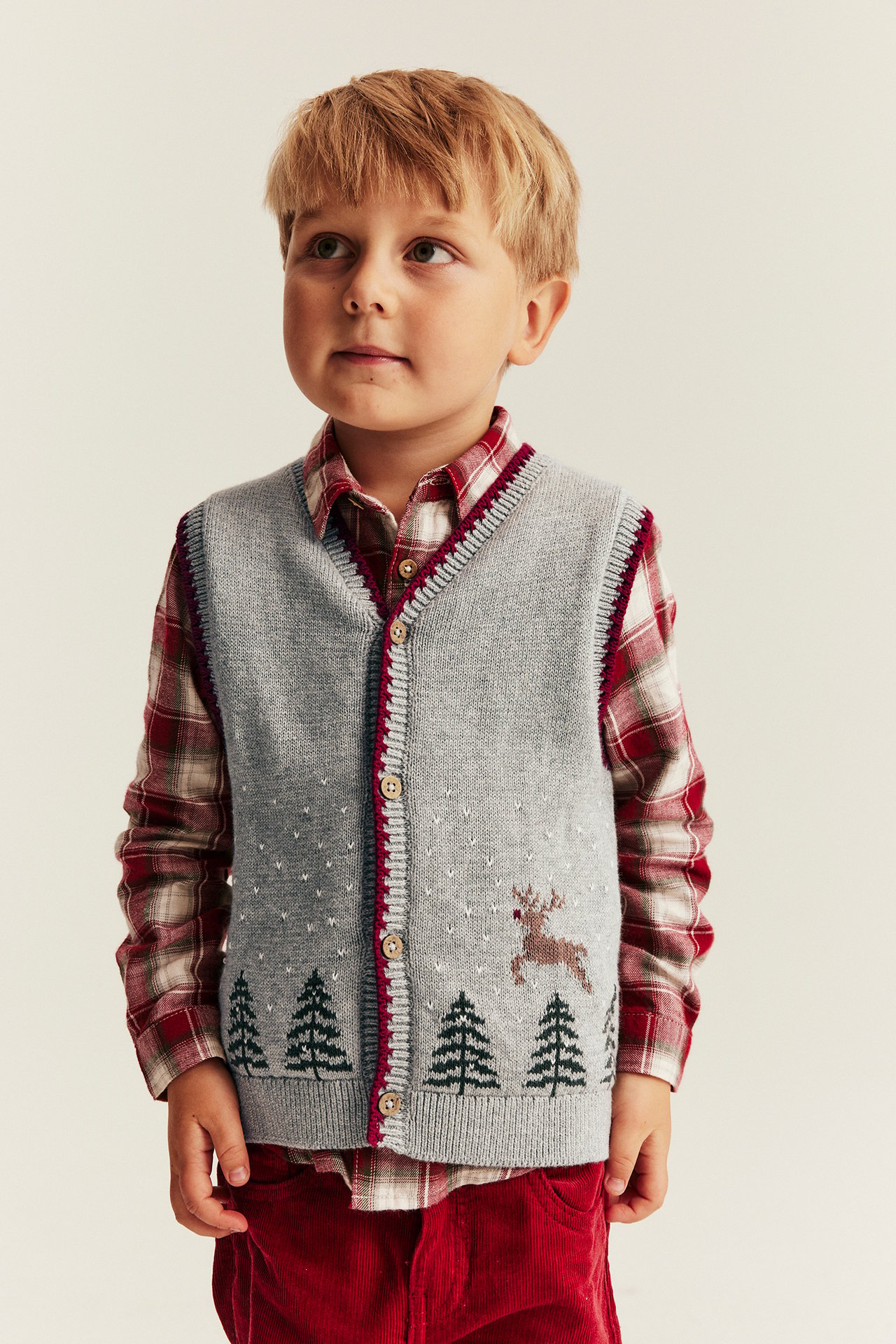 Vorderansicht eines Jungen in grauer gestrickter Weihnachtsweste über einem rot karierten Hemd und roten Cordhosen.