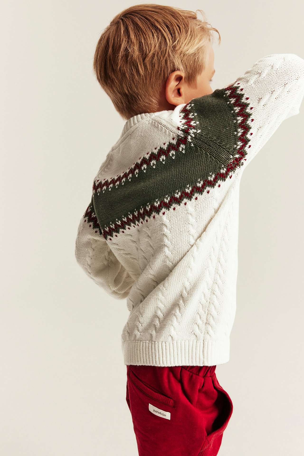 Rückenansicht Kind in cremefarbenem Zopfmusterpullover mit grünem Fair Isle-Muster und roten Jogginghosen.