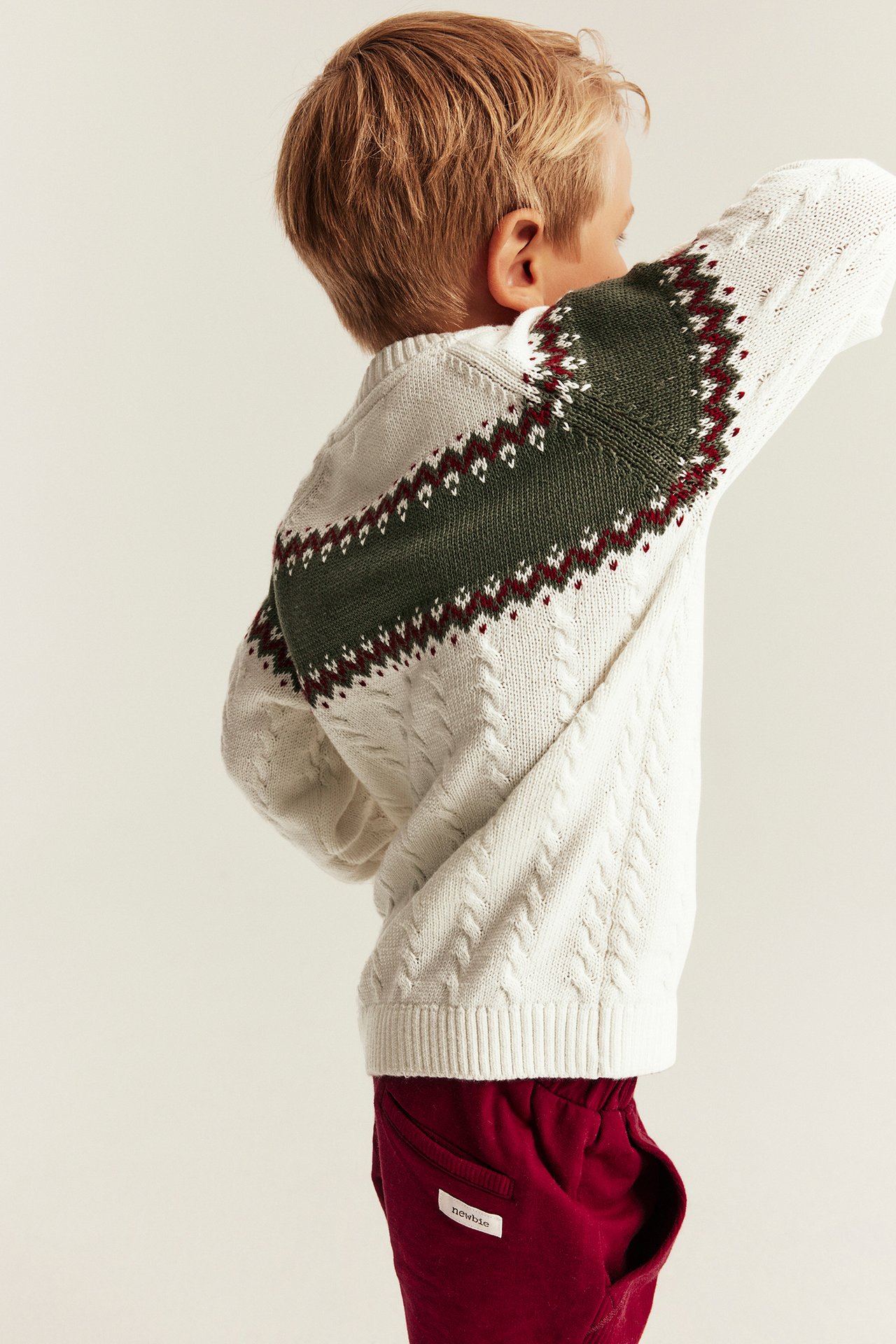 Kind, Rückenansicht: Weißer Zopfstrickpullover mit grünem und rotem Fair Isle-Muster und roter Jogginghose.