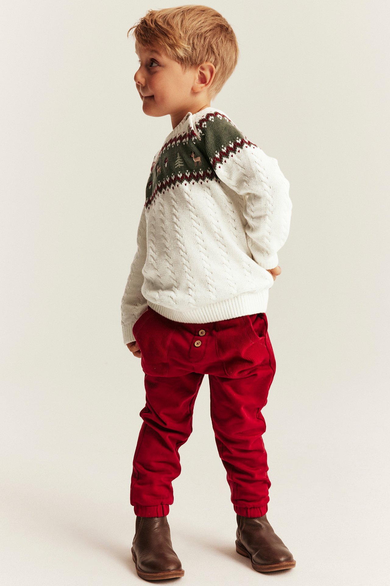 Side view of a kid wearing a white cable knit fair isle sweater, red corduroy pants, and brown boots.