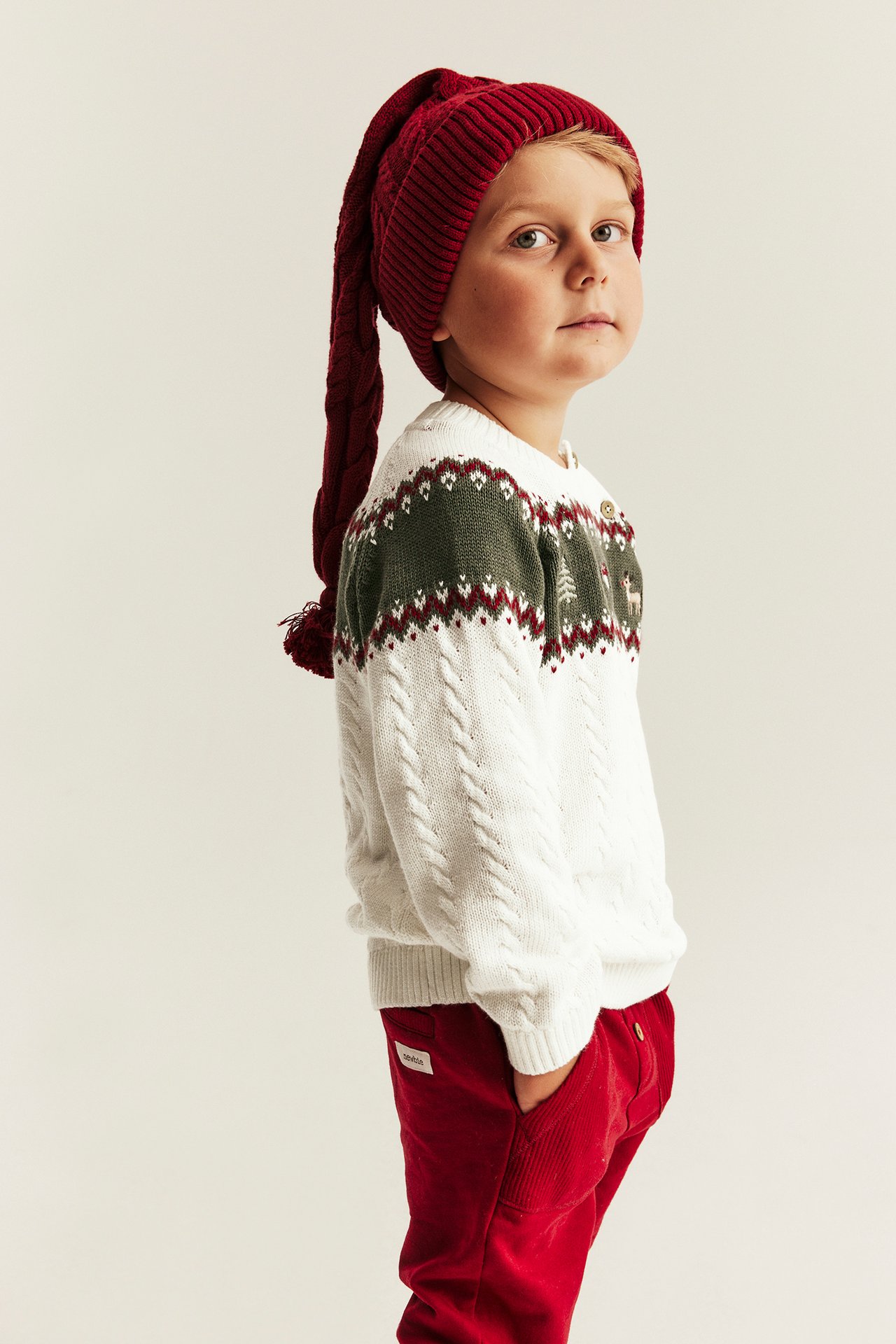 Kid side view: white cable knit fair isle sweater with buttons, long red hat, red corduroy trousers.