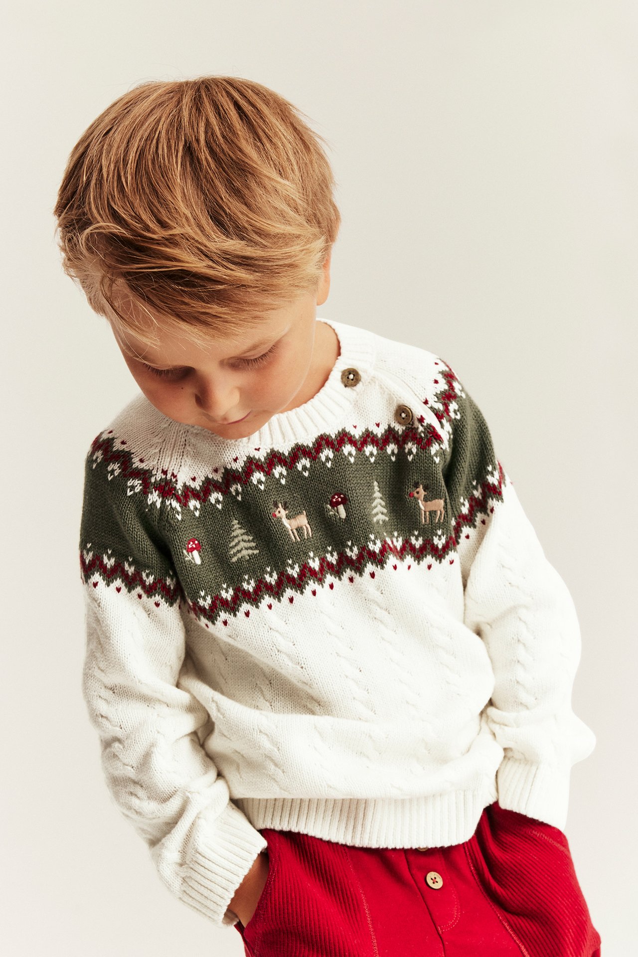 Front view of kid in white and green Fair Isle sweater with deer print, and red corduroy pants.