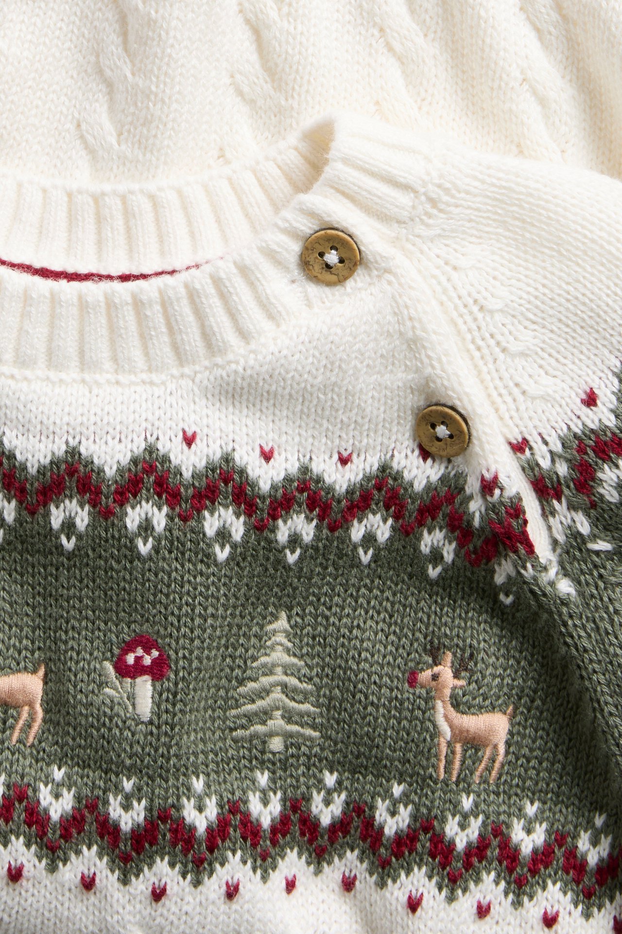 Nahaufnahme eines Baby-Strickpullovers in Creme und Olivgrün mit Fair Isle, Waldbestickung und Holzknöpfen.