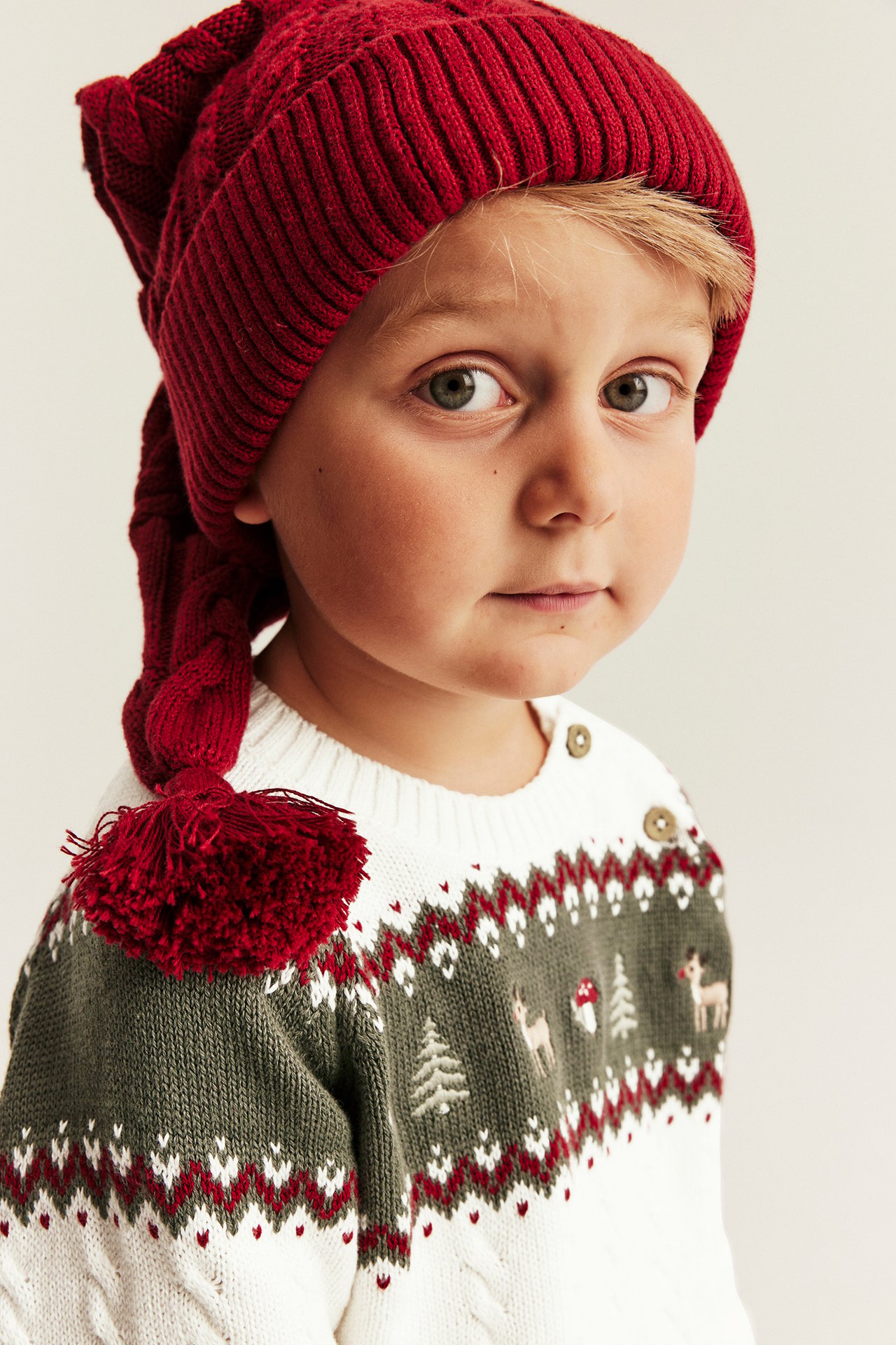 Close-up of a kid wearing a red knitted hat with a pompom and a white and green Christmas sweater.