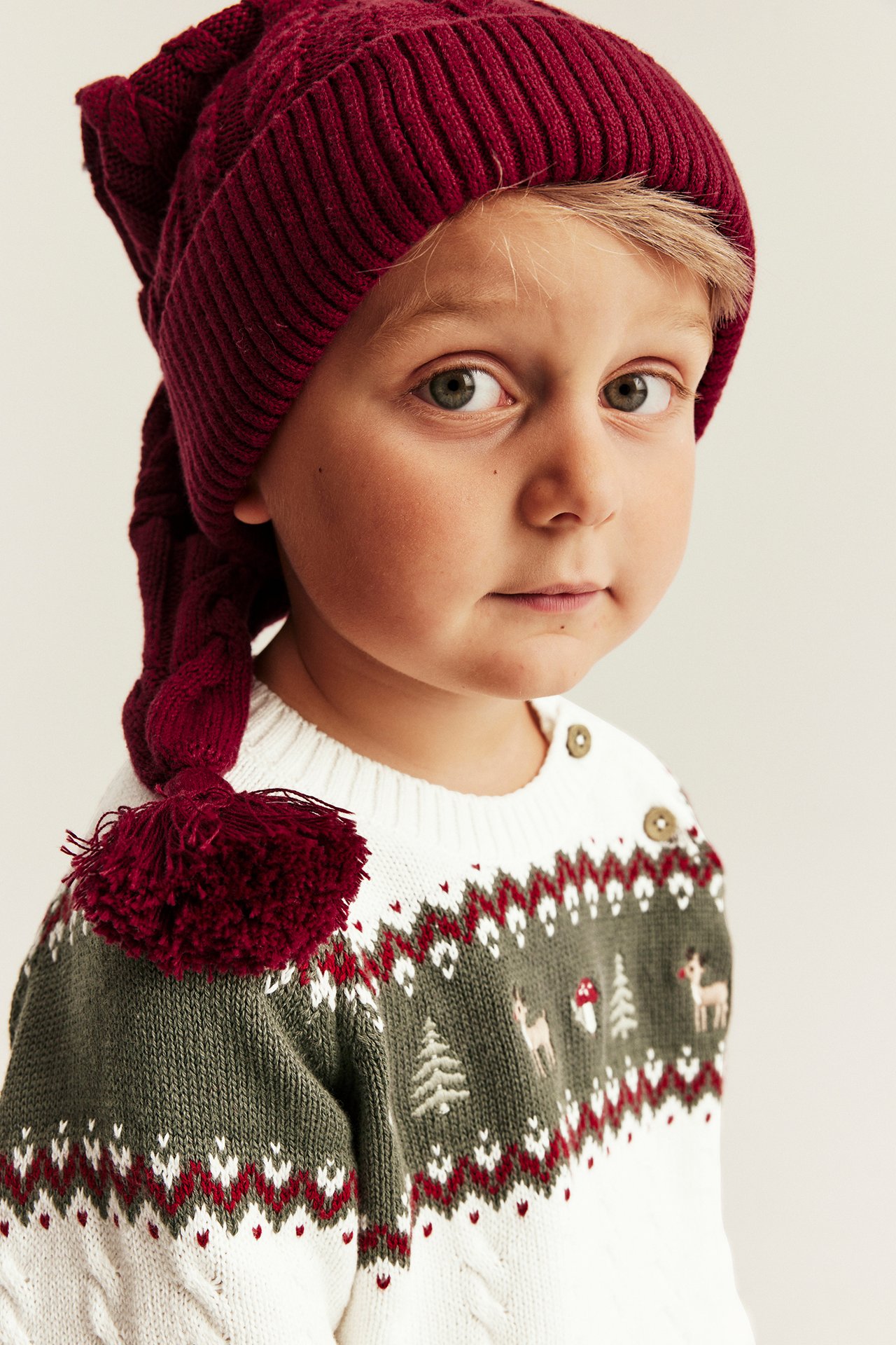 Nahaufnahme: Kind trägt rote Strickmütze mit Zopfgeflecht, Bommel und weiß-grünen Weihnachtspullover.
