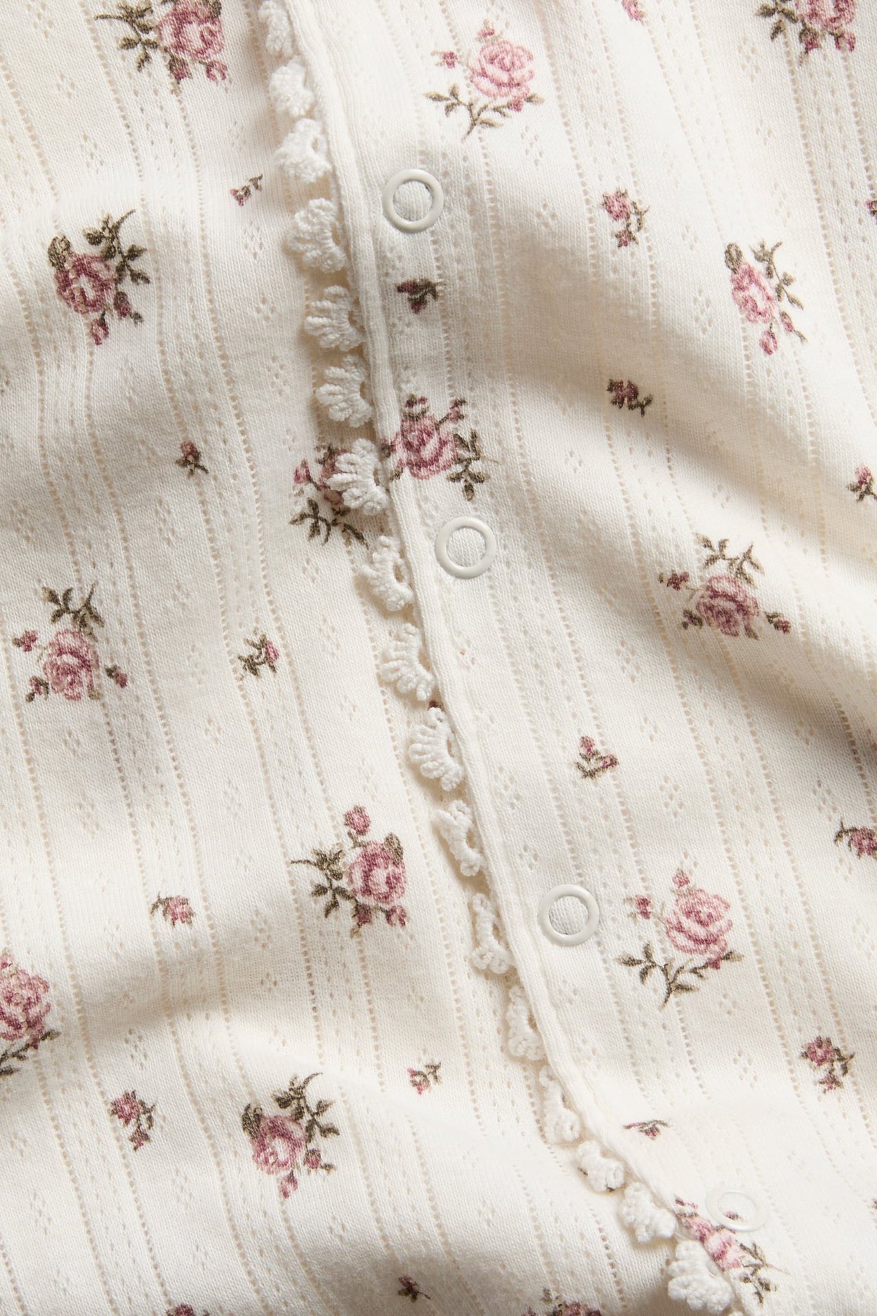 Close-up of a cream ribbed knit garment with pink rose print, white snap buttons, and ruffle trim.