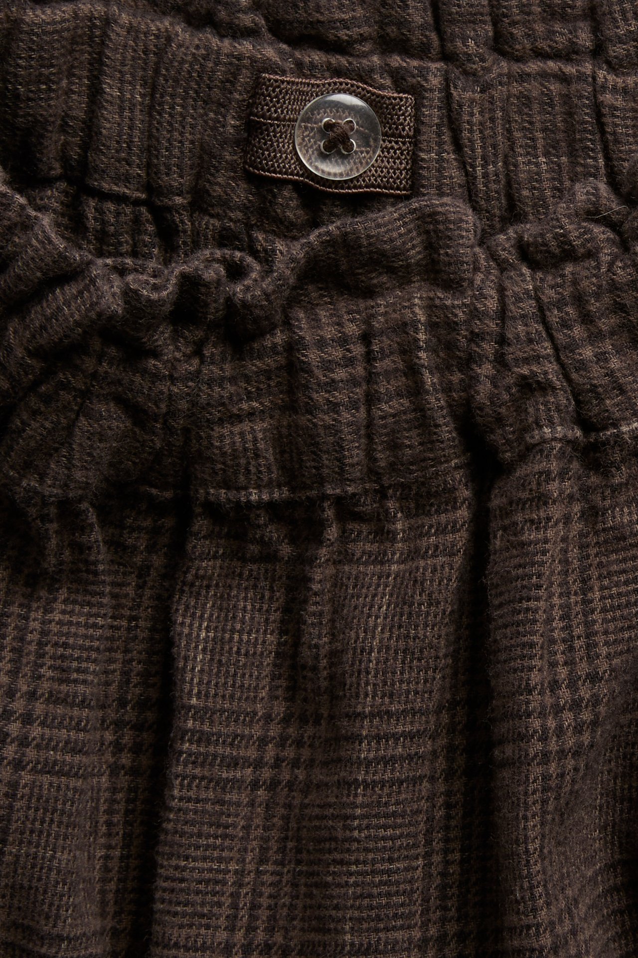 Close-up of dark brown plaid garment fabric with an elasticized section and a clear button.