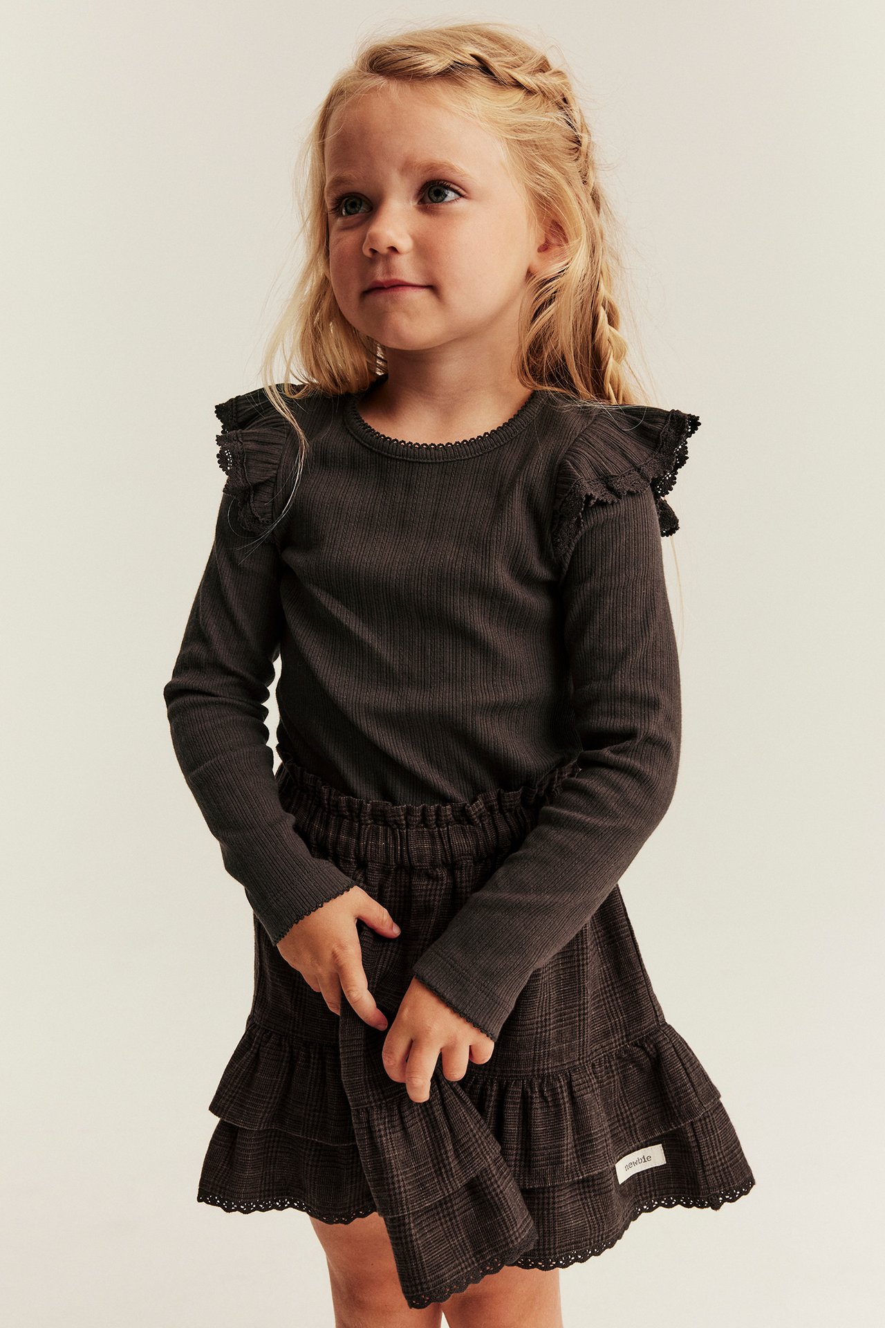 Front view of a kid wearing a dark ribbed long-sleeve top with ruffles and a plaid tiered skirt.