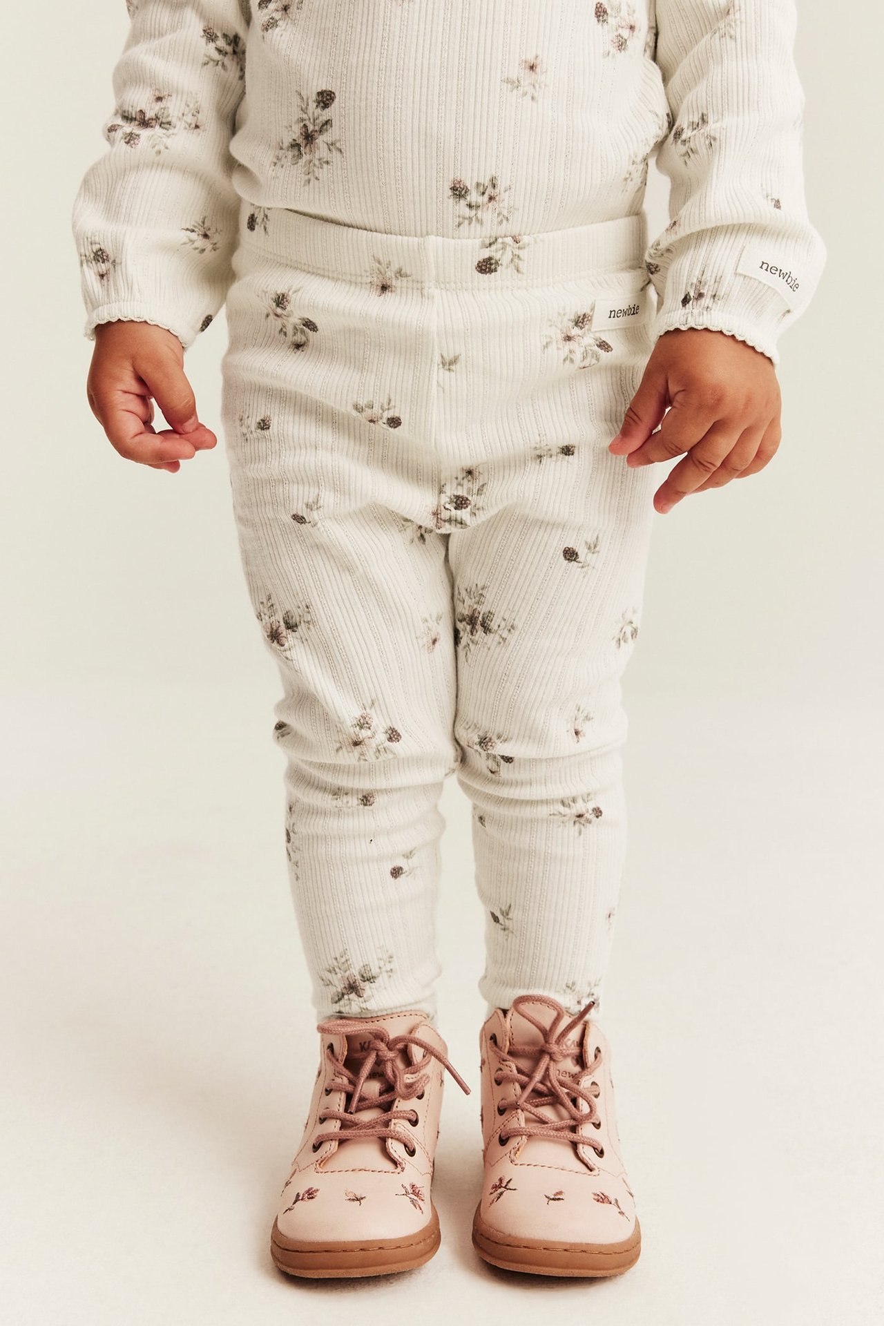 Vorderansicht, Baby trägt cremefarbenen gerippten Pyjama mit Blumendruck und rosa Schnürstiefel.