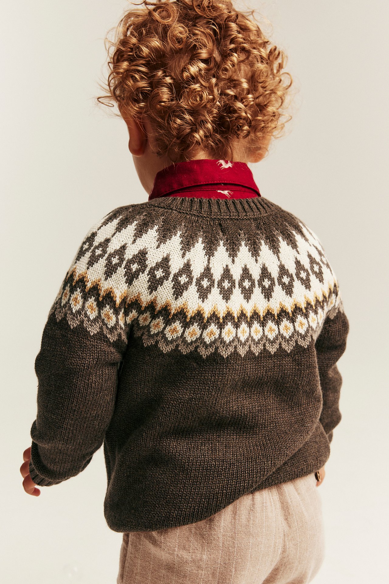 Back view of kid in dark brown patterned sweater, red horse shirt, light pink pants.