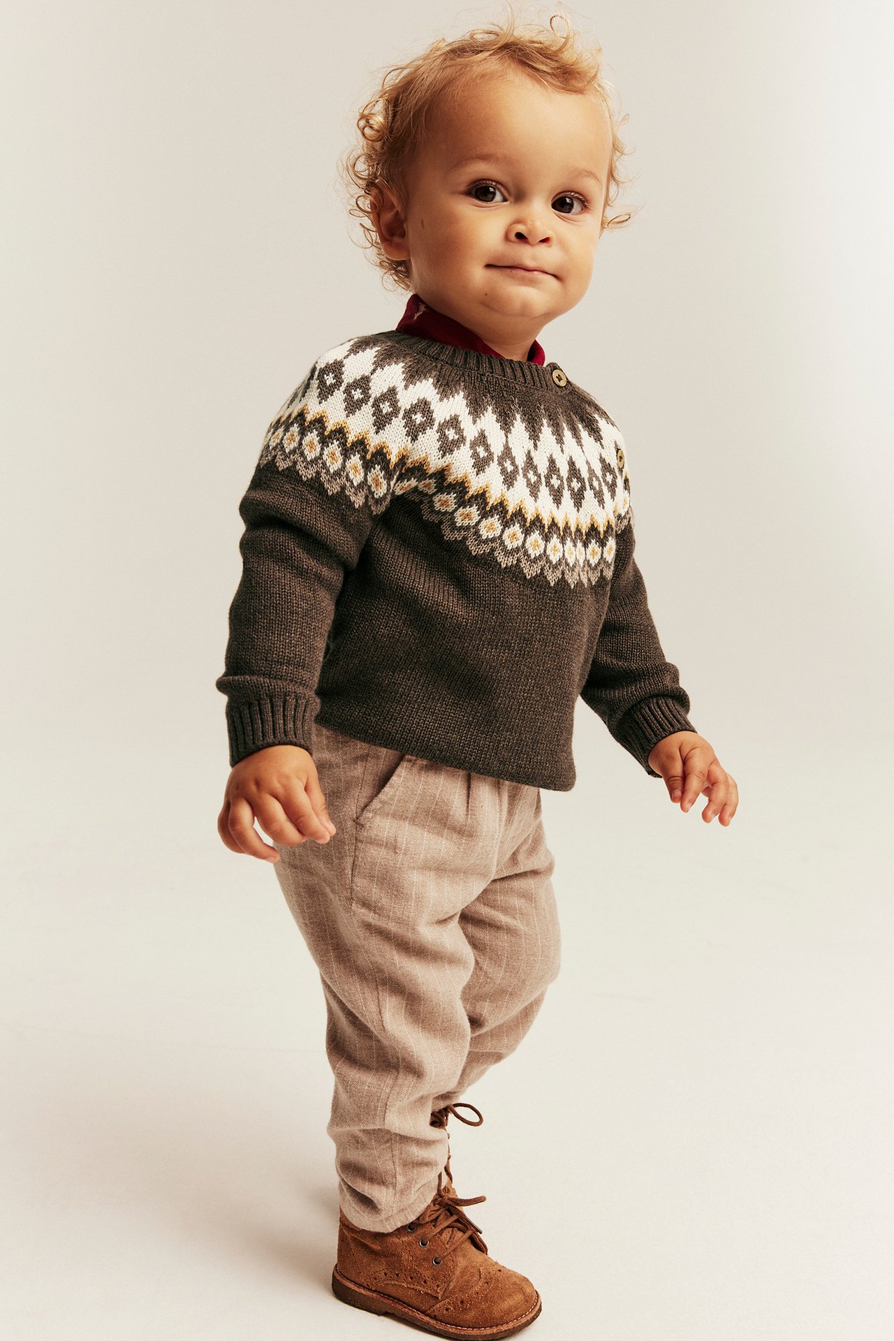 Baby wearing a dark brown fair isle sweater, red shirt, beige pants, and brown boots.