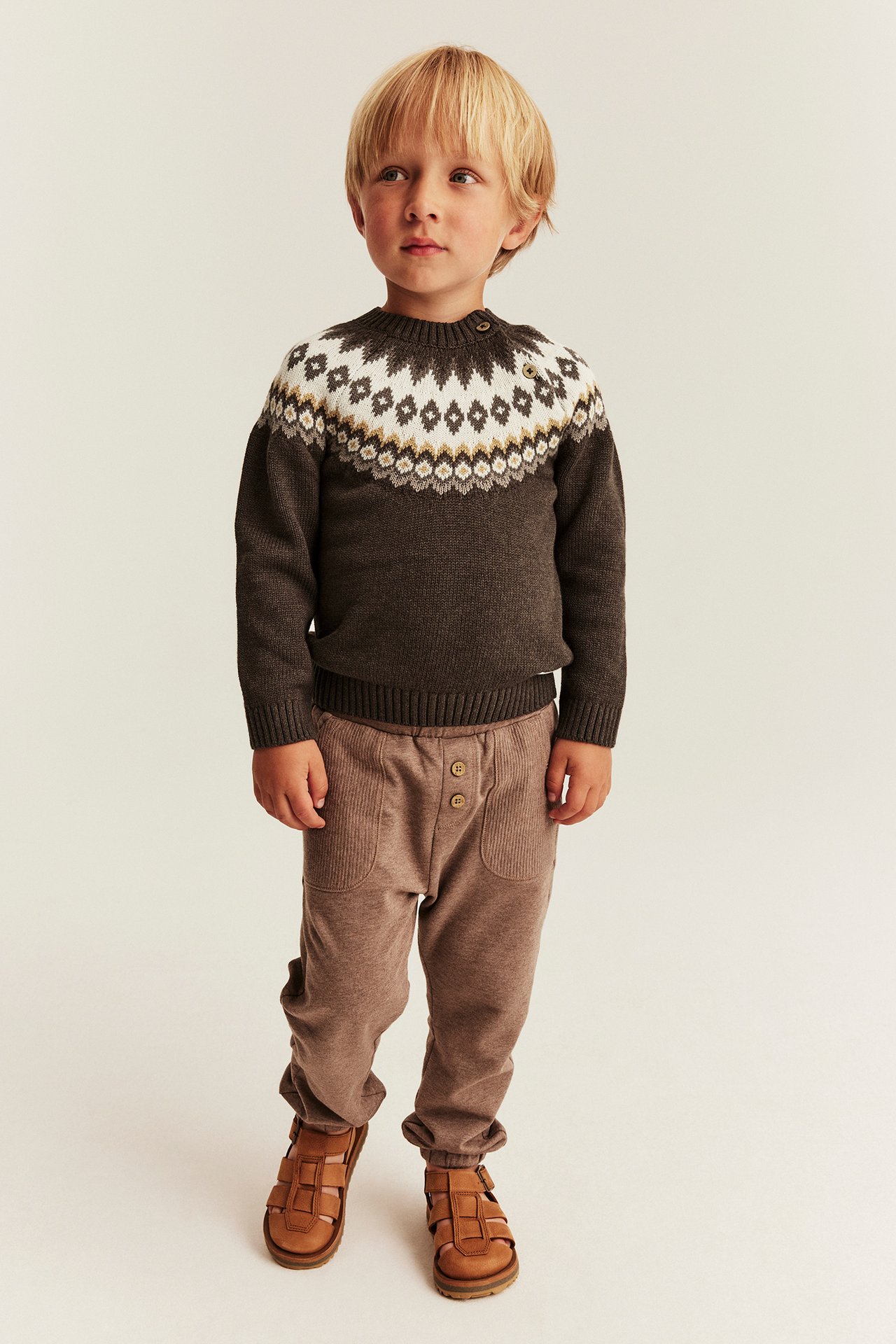 Vorderansicht: Kind in dunkelbraunem Fair-Isle-Pullover, brauner Ripphose und braunen Sandalen.