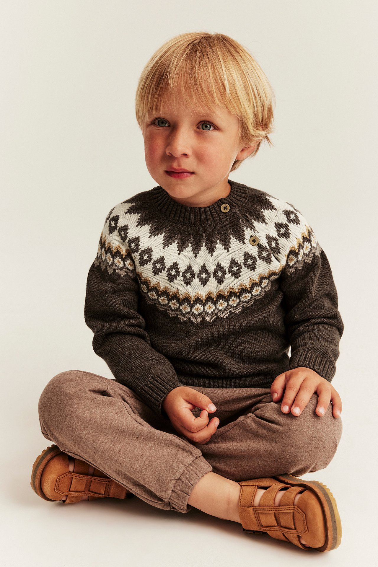 Vorderansicht eines Kindes in braunem Fair-Isle-Pullover, brauner Jogginghose und braunen Sandalen.