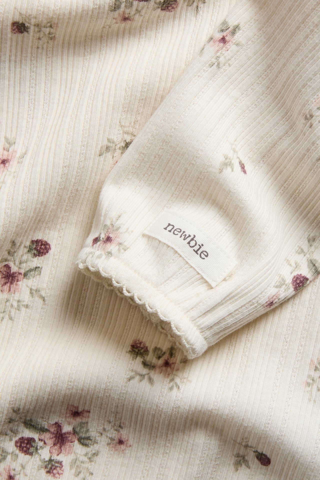 Close-up of an off-white ribbed baby garment with purple floral print and 'newbie' label on sleeve.