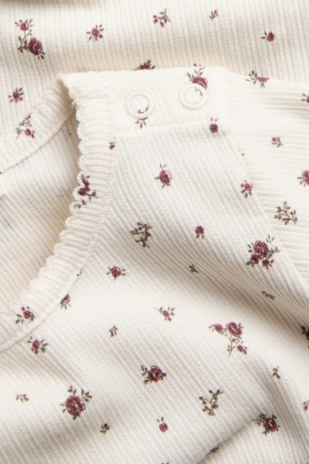 Nahaufnahme eines cremefarbenen Baby-Bodys aus Rippstrick mit kleinen dunkelroten Blumen und Druckknöpfen.