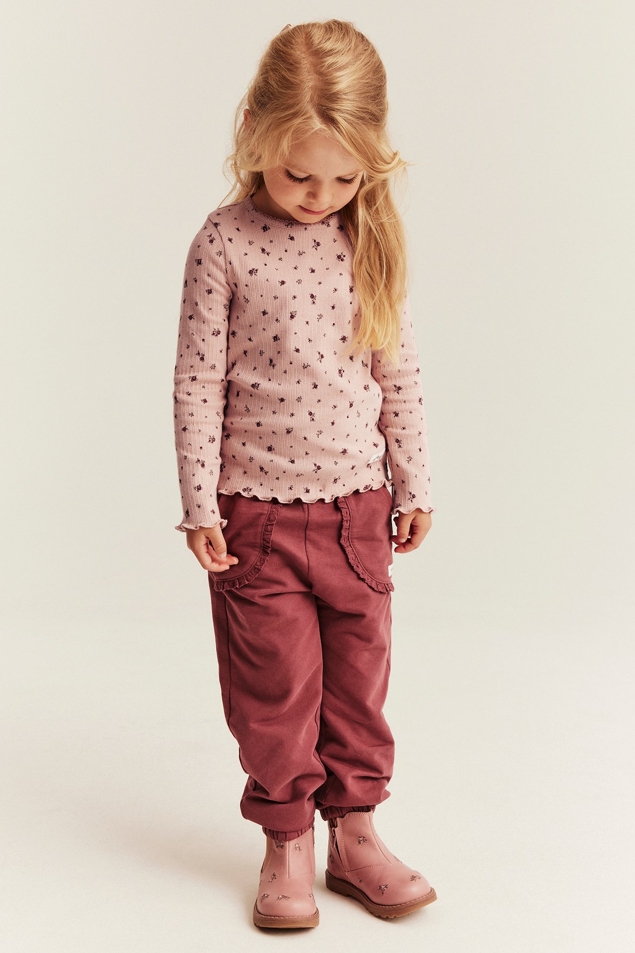 Vorderansicht, Kind in rosa geblümtem geripptem Langarmshirt, weinroter Jogginghose und rosa bestickten Stiefeln.