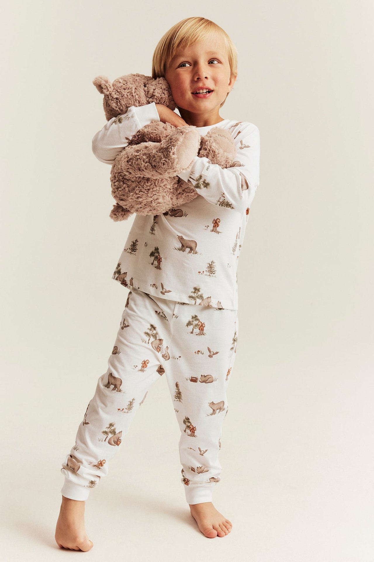 Vorderansicht: Kind in weißem Schlafanzug mit Waldtiermuster, hält einen braunen Teddybären.