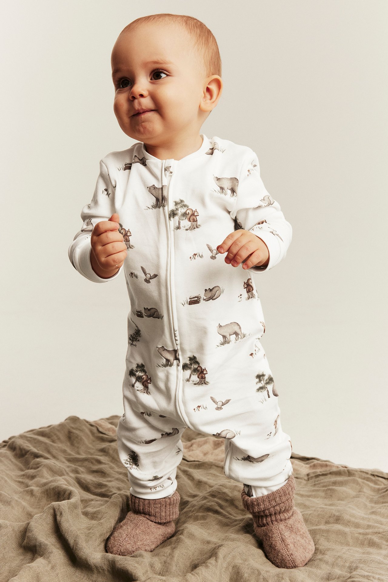 Front view of a baby wearing a white long-sleeved full-zip animal print jumpsuit with brown socks.