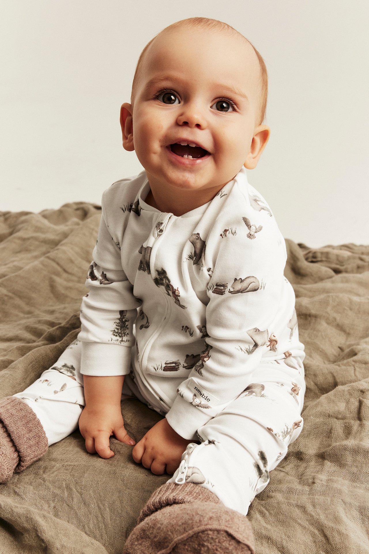 Front view of a smiling baby wearing a white animal print long-sleeved romper with a zipper and brown knitted socks.