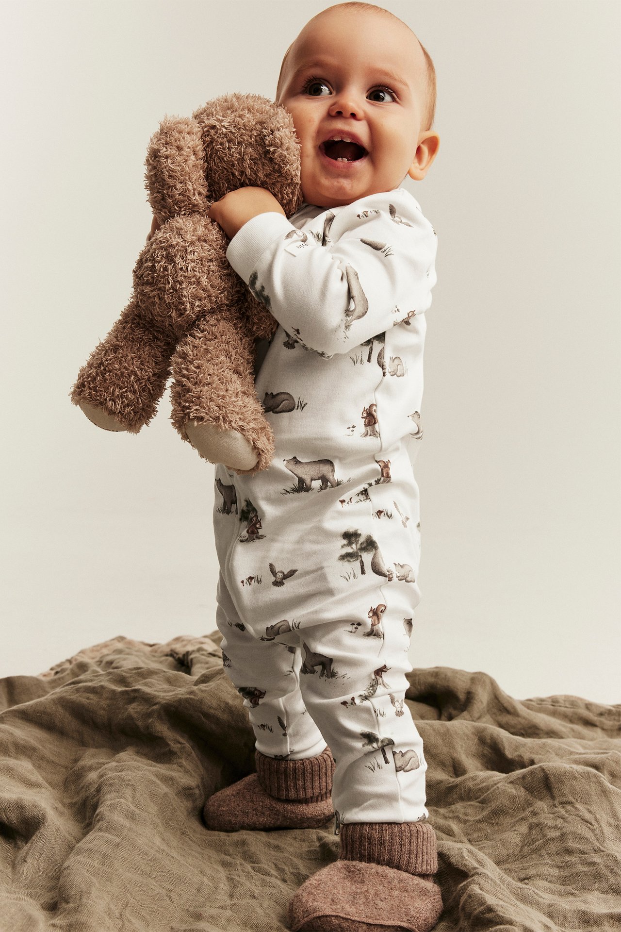 Front view: Baby in white animal print jumpsuit, holding brown teddy bear and wearing brown slippers.