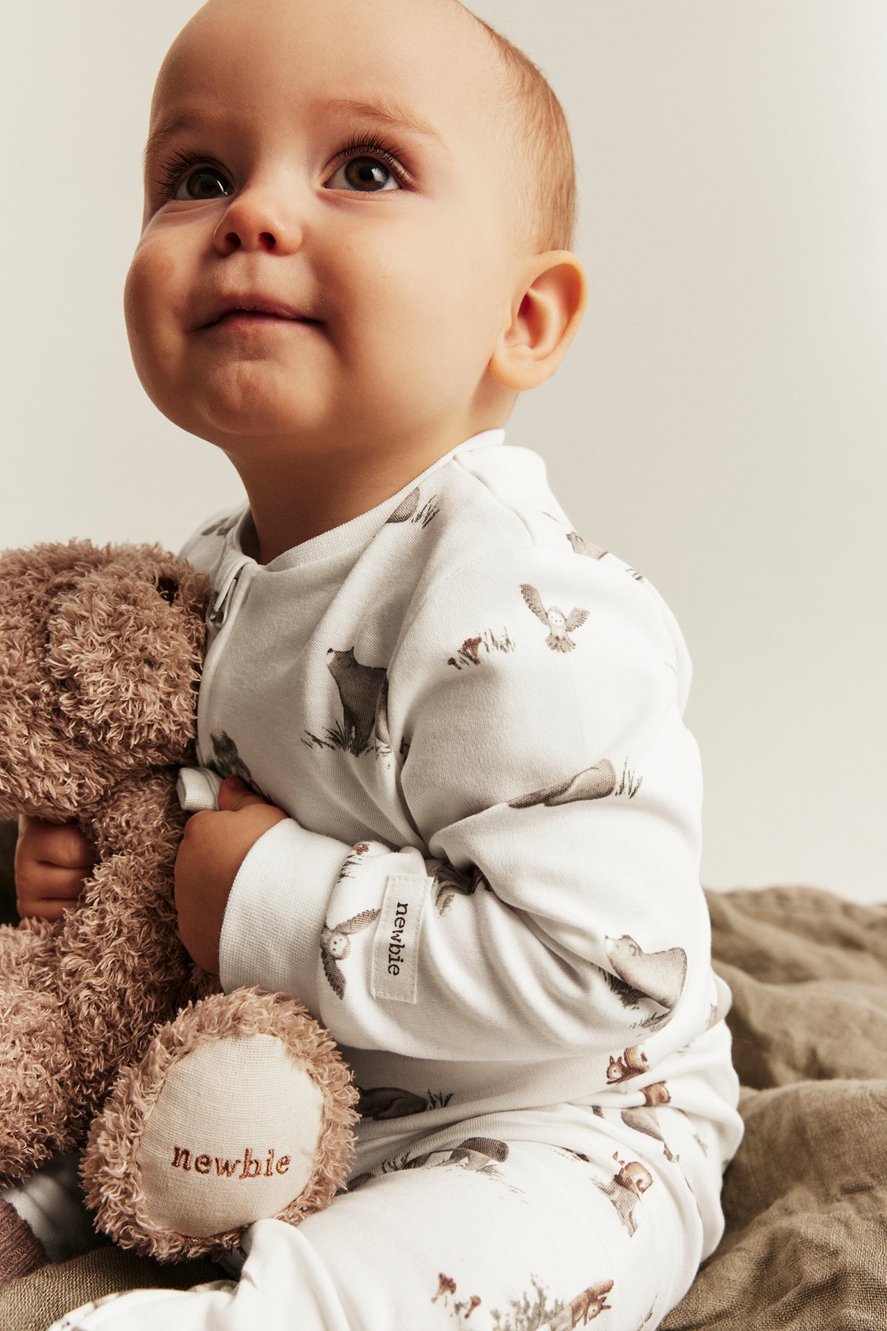 Close-up of a baby wearing a white long-sleeved bodysuit with forest animal print, holding a brown teddy bear.