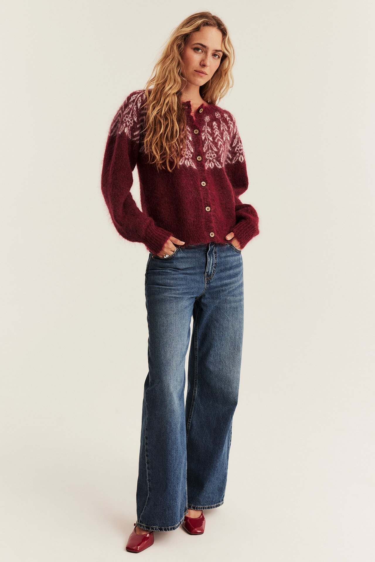 Vorderansicht: Frau trägt bordeauxroten gemusterten Strickcardigan, weite blaue Jeans, rote Lackschuhe.