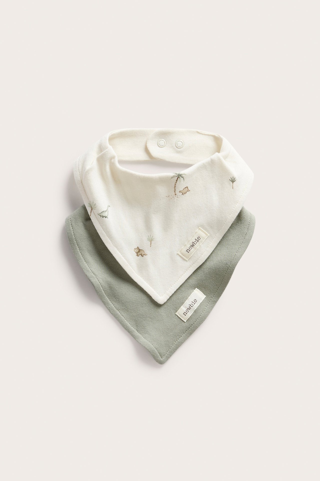 Front view of two baby bibs: one off-white with dinosaur print, one solid sage green, both with snap fastenings.