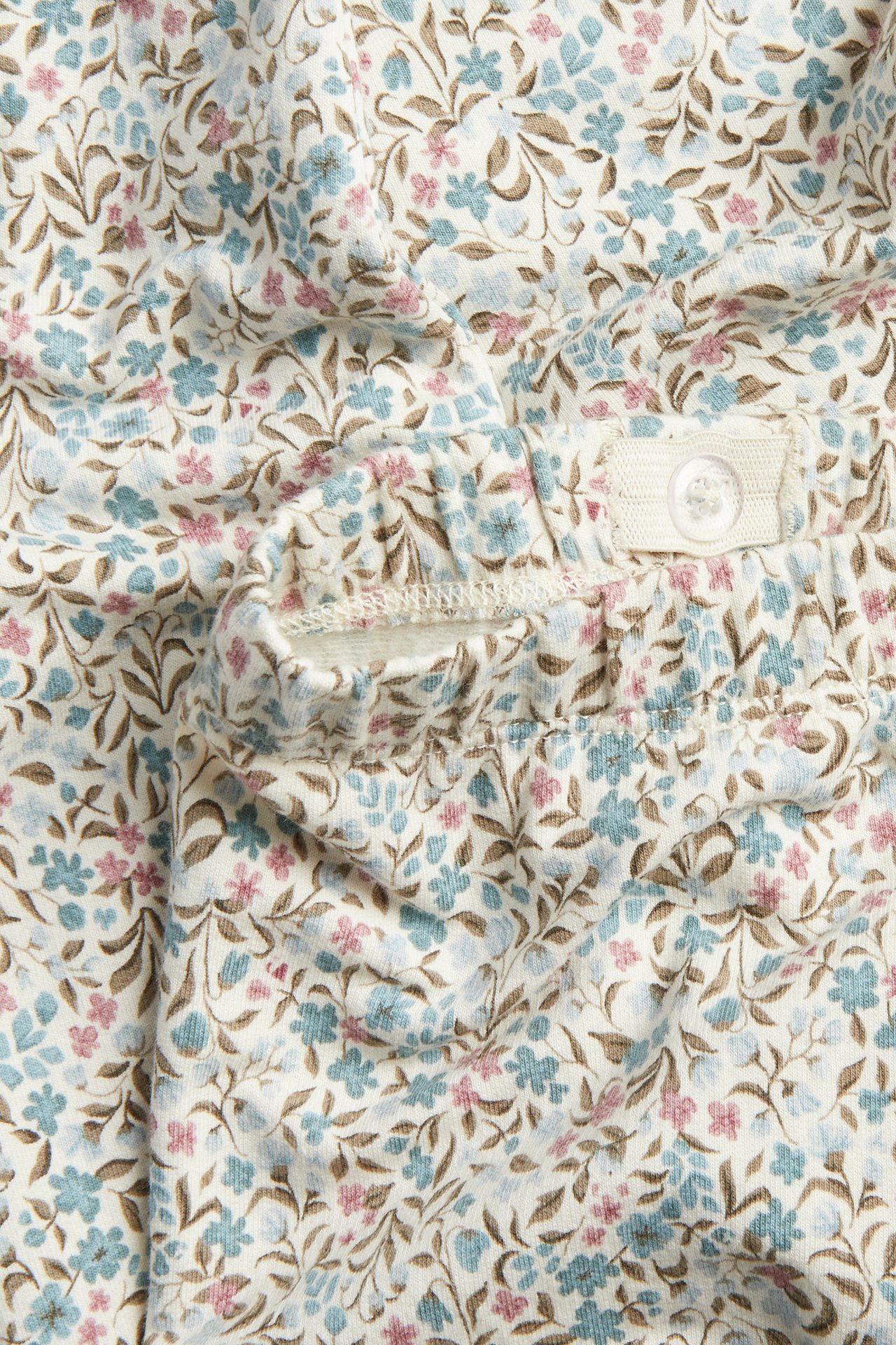 Nahaufnahme: Weißes Blumenmuster-Kleidungsstück mit blauen, rosa, braunen Blüten und Knopf am Bund.