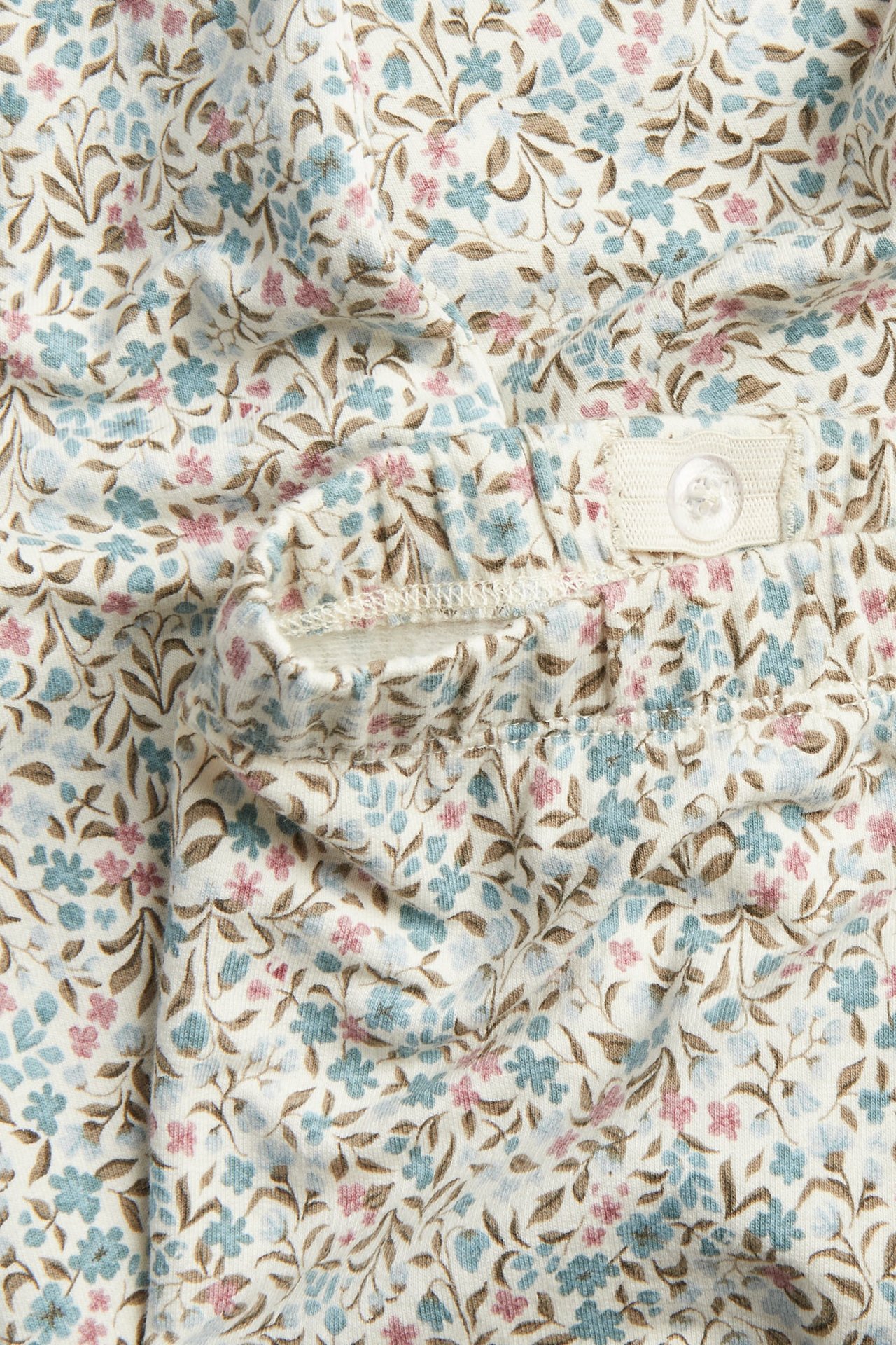 Nahaufnahme: Weißes Blumenmuster-Kleidungsstück mit blauen, rosa, braunen Blüten und Knopf am Bund.