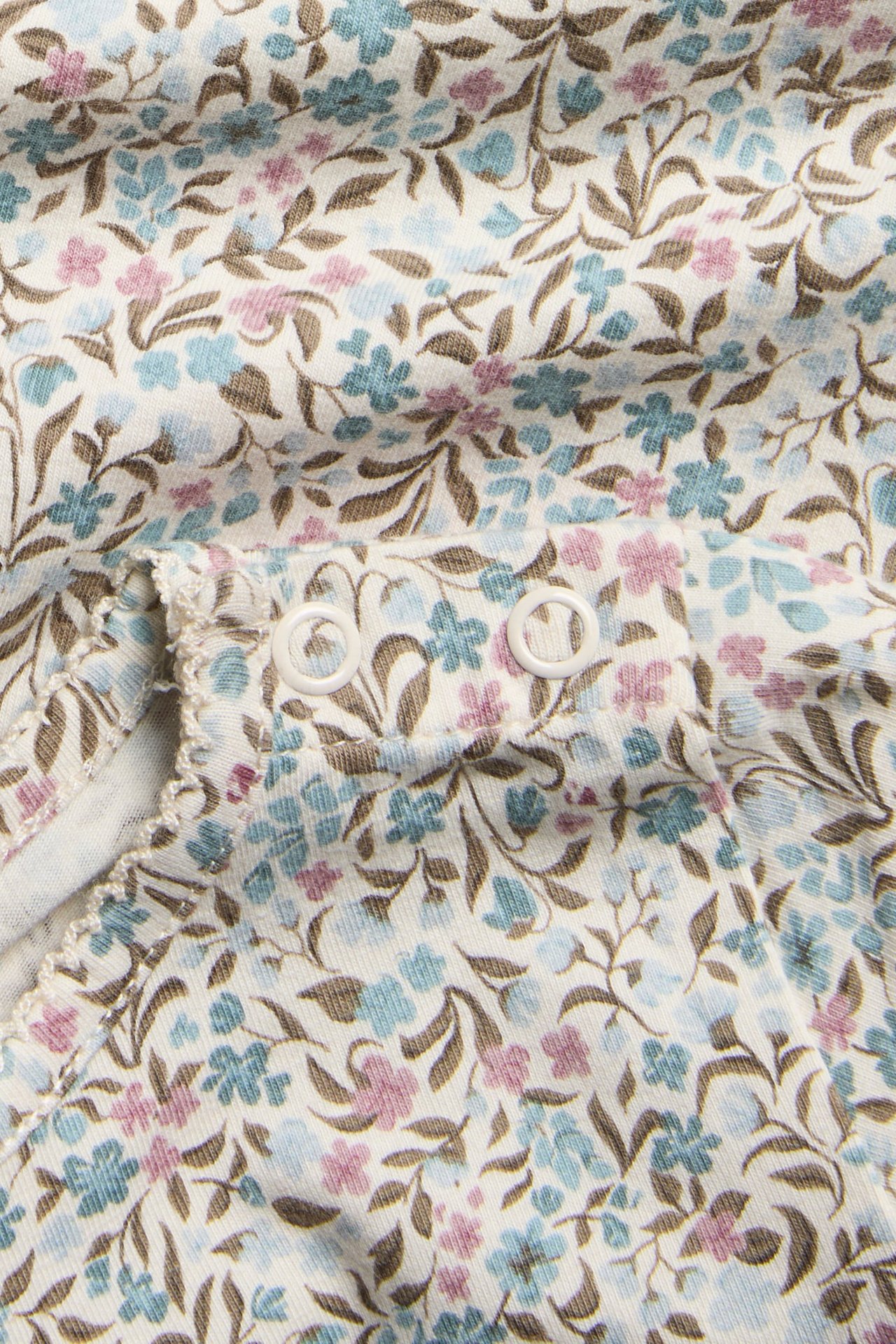 Nahaufnahme eines Babykleidungsstücks mit cremefarbenem Blumenmuster, blauen/rosa Blüten und weißen Druckknöpfen.