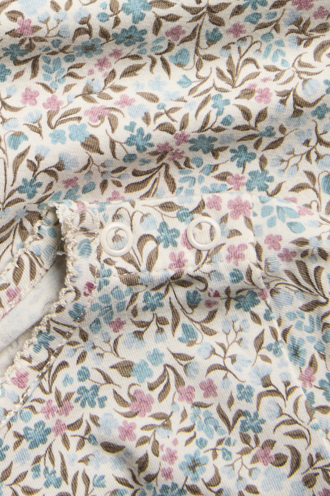 Nahaufnahme eines Babykleidungsstücks mit cremefarbenem Blumenmuster, blauen/rosa Blüten und weißen Druckknöpfen.