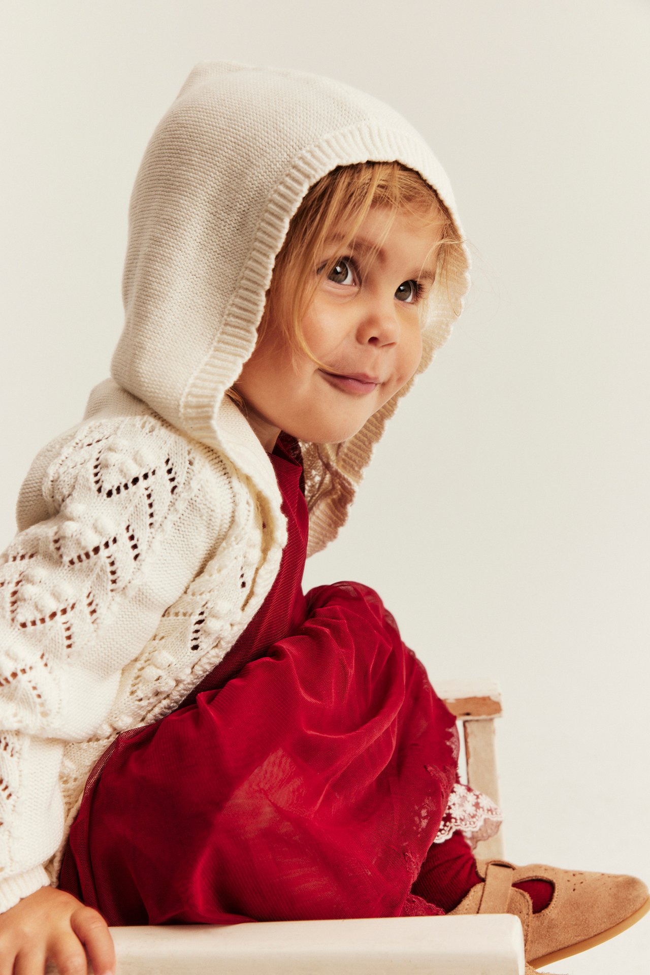 Nahaufnahme eines Kindes in cremefarbener Strickjacke mit Kapuze über rotem Kleid und braunen Schuhen.