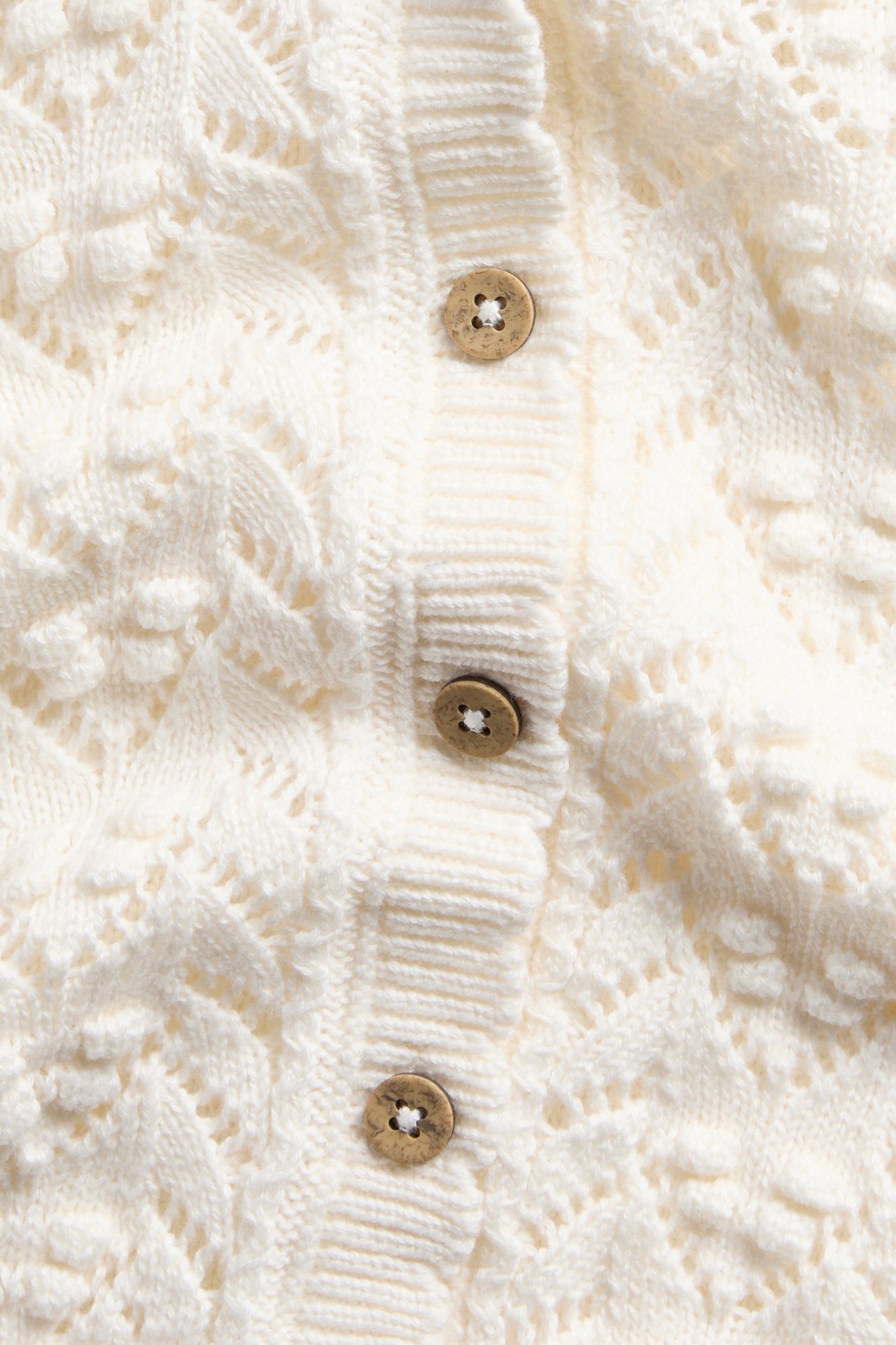 Nahaufnahme eines cremefarbenen strukturierten Strickcardigans mit Lochmuster und drei Holzknöpfen.