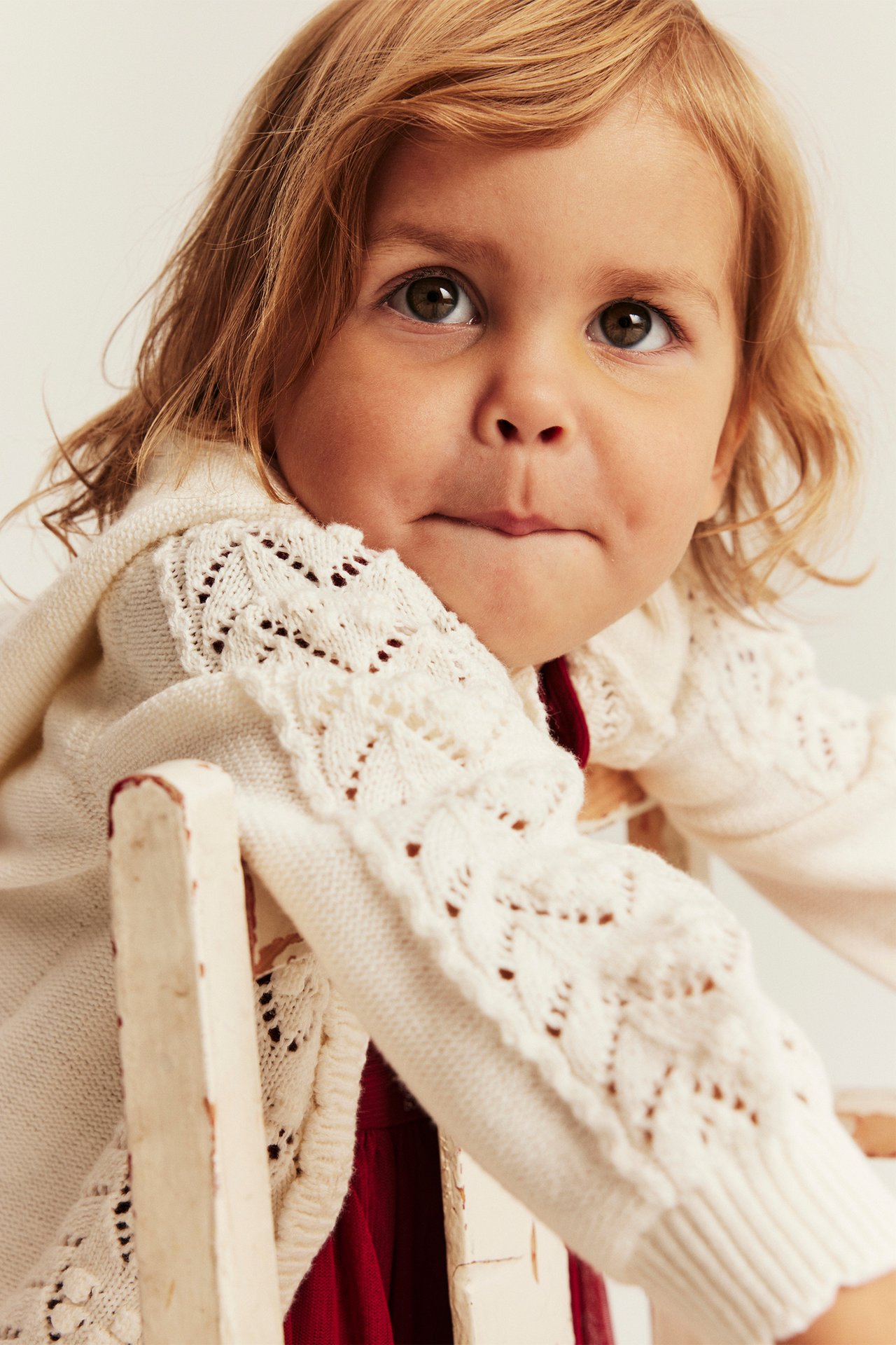 Nahaufnahme eines Kindes in einer cremefarbenen Strickjacke mit Lochmuster über einem roten Oberteil.