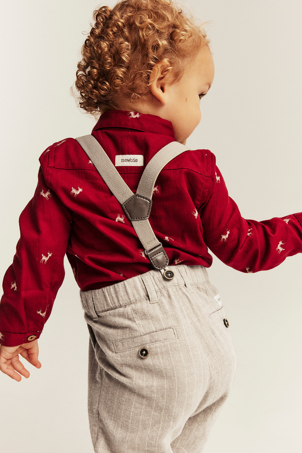 Rückansicht eines Babys in rotem Hemd mit Rentierdruck, heller Nadelstreifenhose und grauen Hosenträgern.