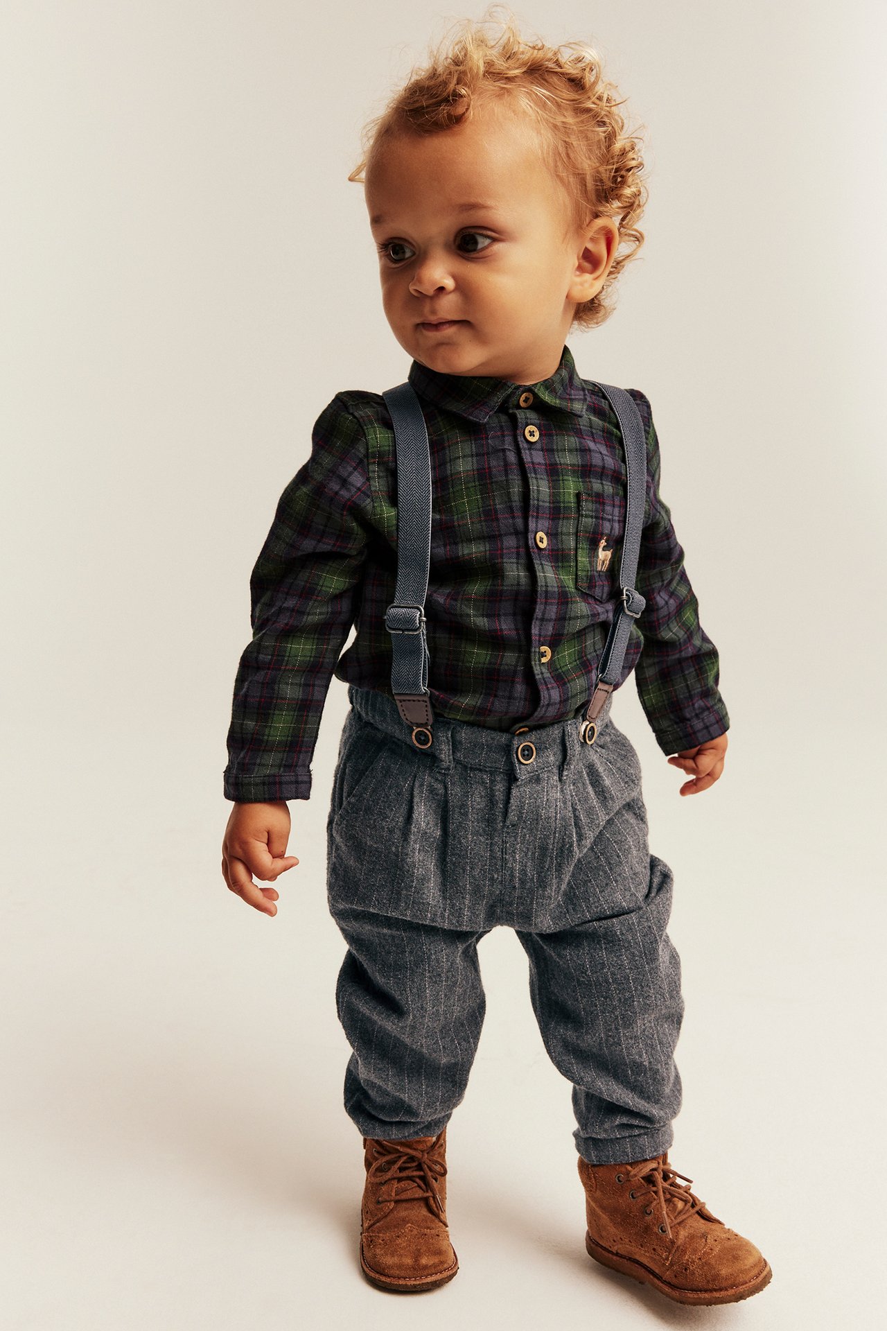 Front view of a baby boy in a green plaid shirt, gray pinstripe suspender pants, and brown boots.