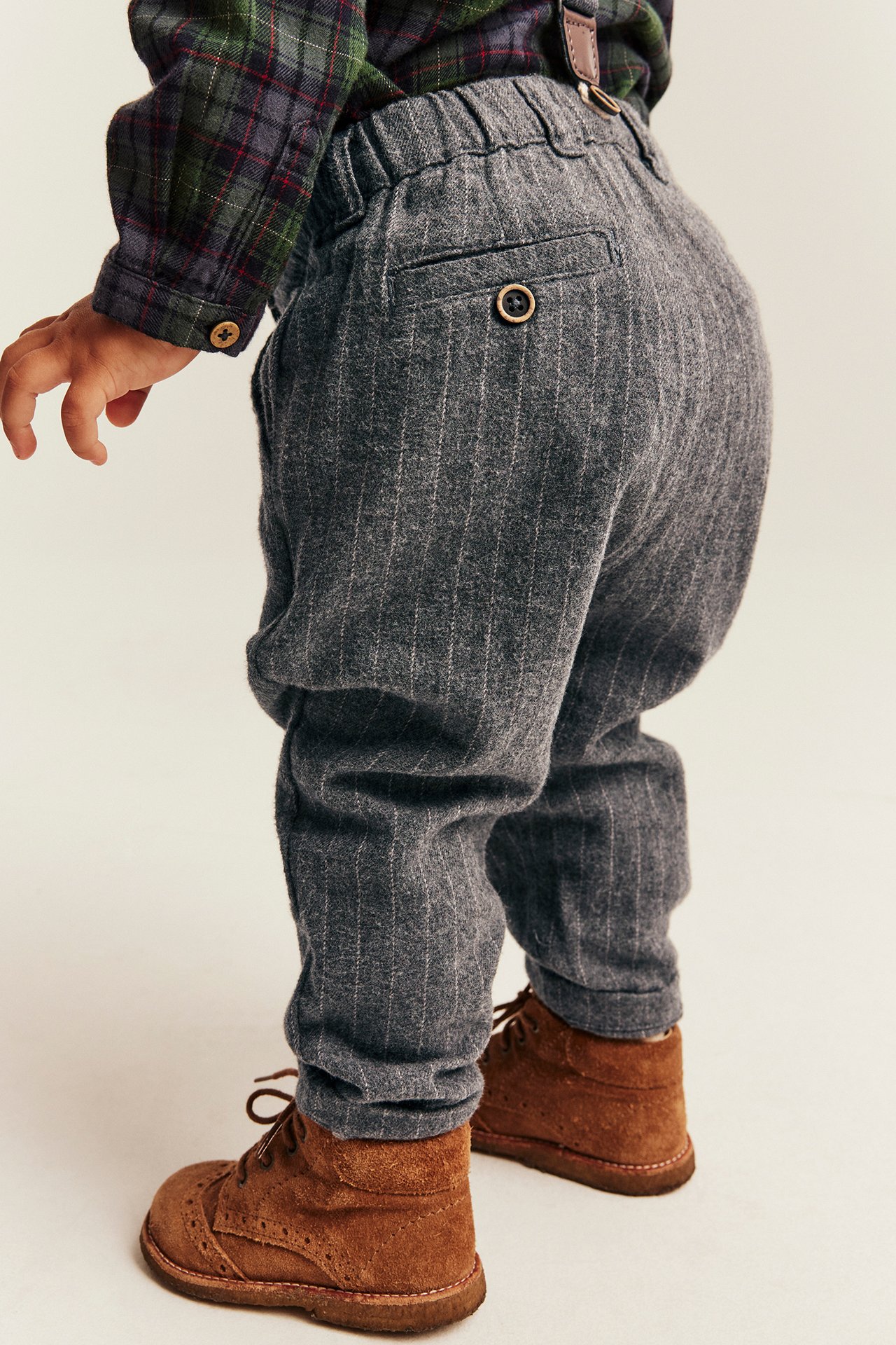 Back view of baby in grey pinstripe trousers, plaid shirt, brown suspenders, and brown boots.