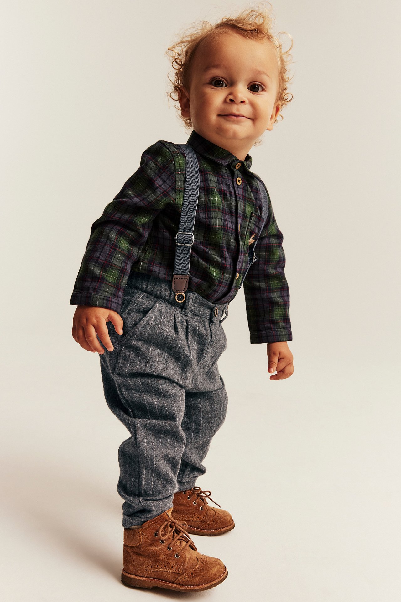 Baby in grün-kariertem Hemd, grau gestreiften Hosen mit Hosenträgern und braunen Stiefeln, Vorderansicht.