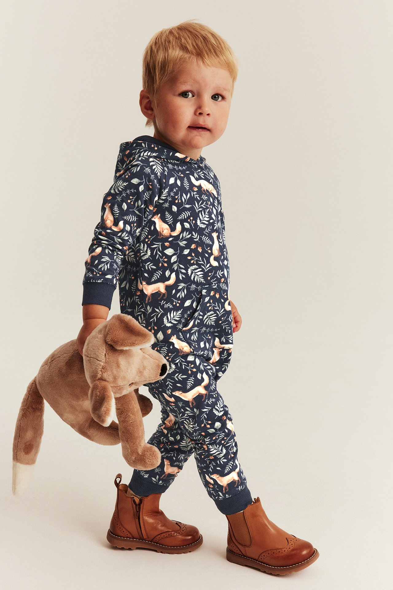 Kid wears dark blue fox print hoodie and sweatpants set, brown boots, holding a brown dog toy.