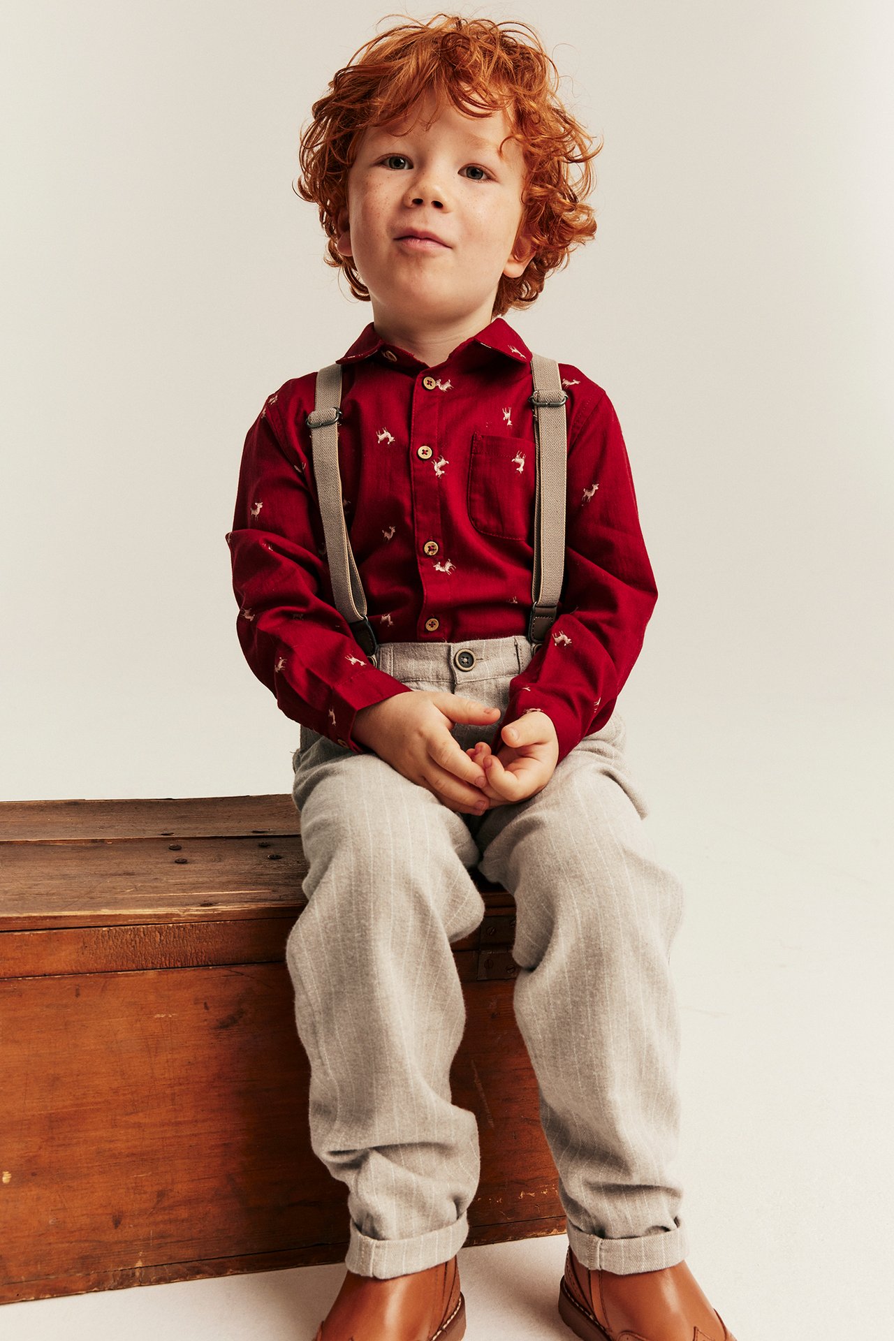 Front view: Kid in red deer print shirt, grey suspenders, light grey pinstripe pants, brown boots.