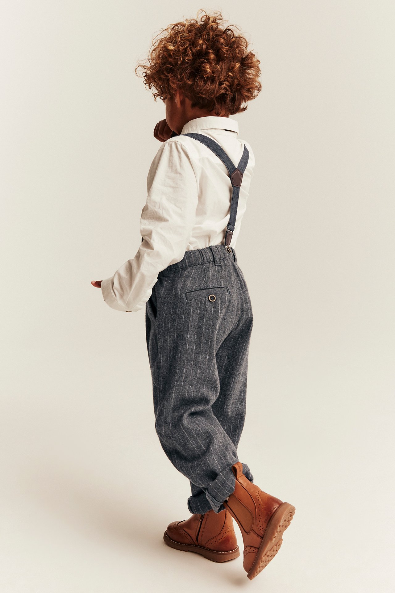 Rückenansicht eines Kindes: weißes Hemd, grau gestreifte Hose, blaue Hosenträger und braune Stiefel.