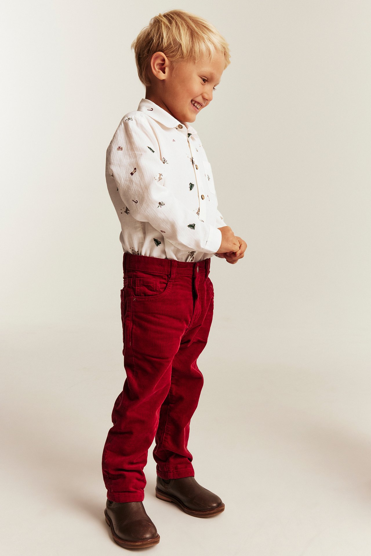 Side view of a kid wearing red corduroy pants, a white nature print shirt, and brown shoes.