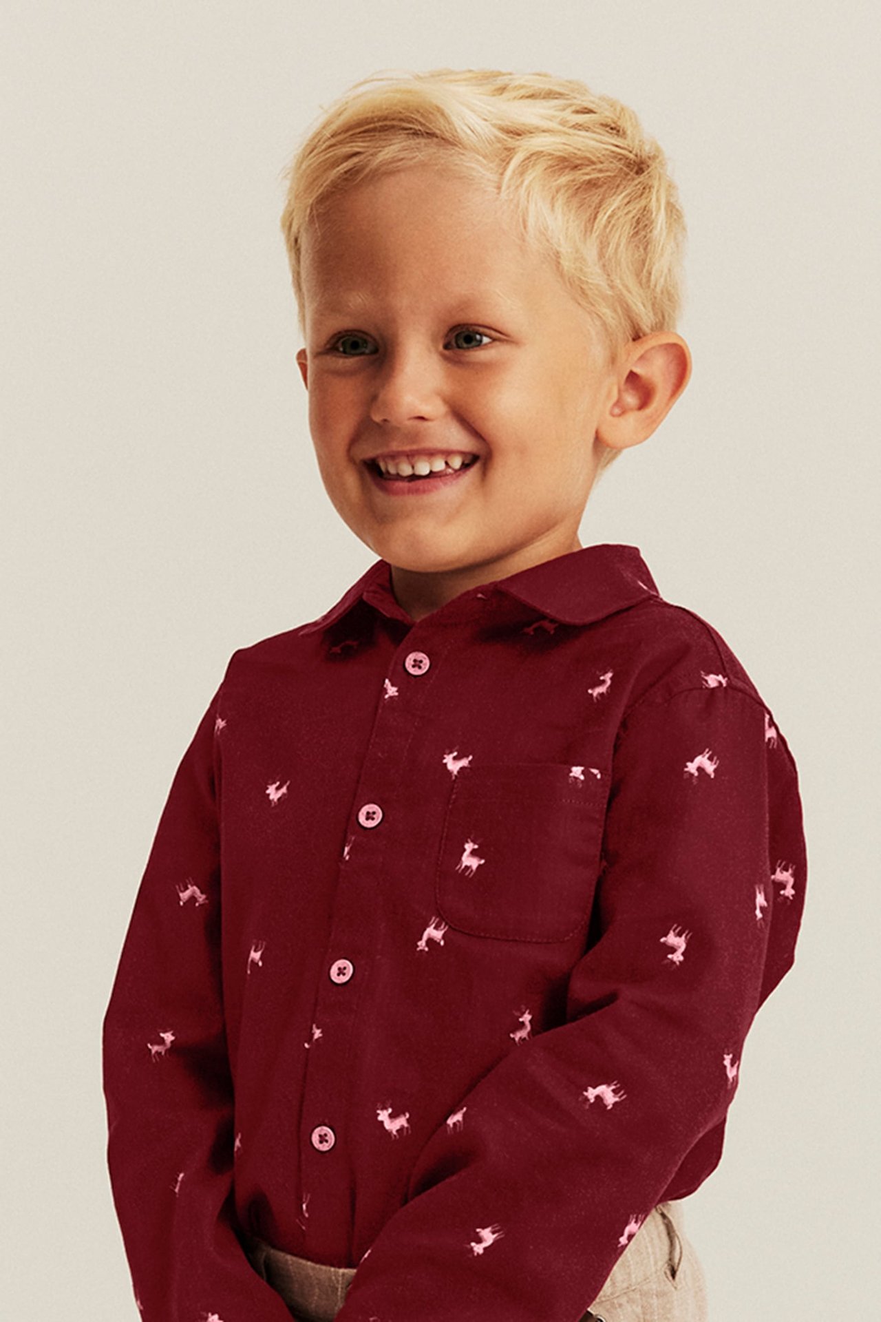 Front view of a kid in a burgundy long-sleeved shirt with pink deer print and beige pants.