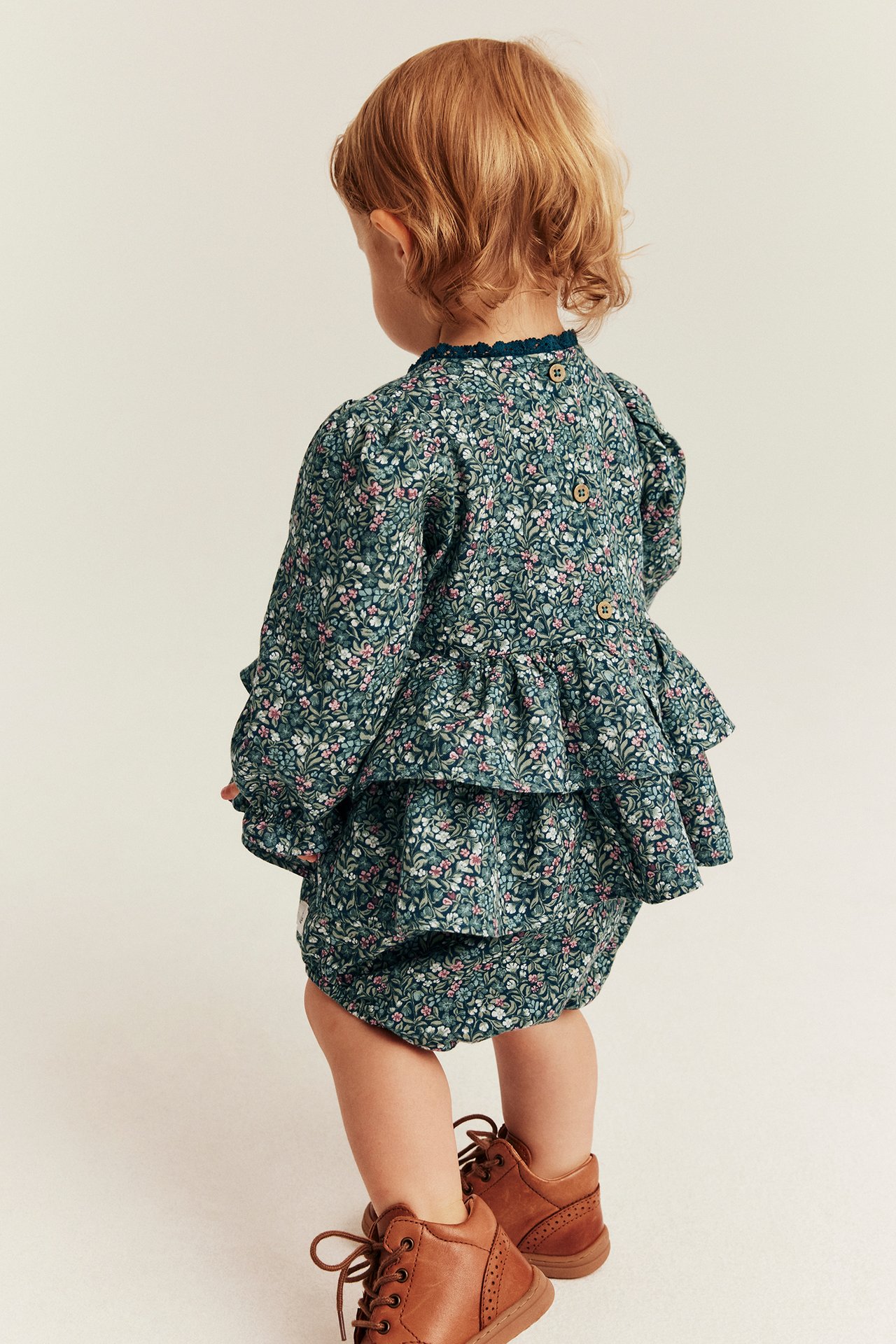 Back view: baby in green floral print top with ruffles, matching bloomers, and brown boots.