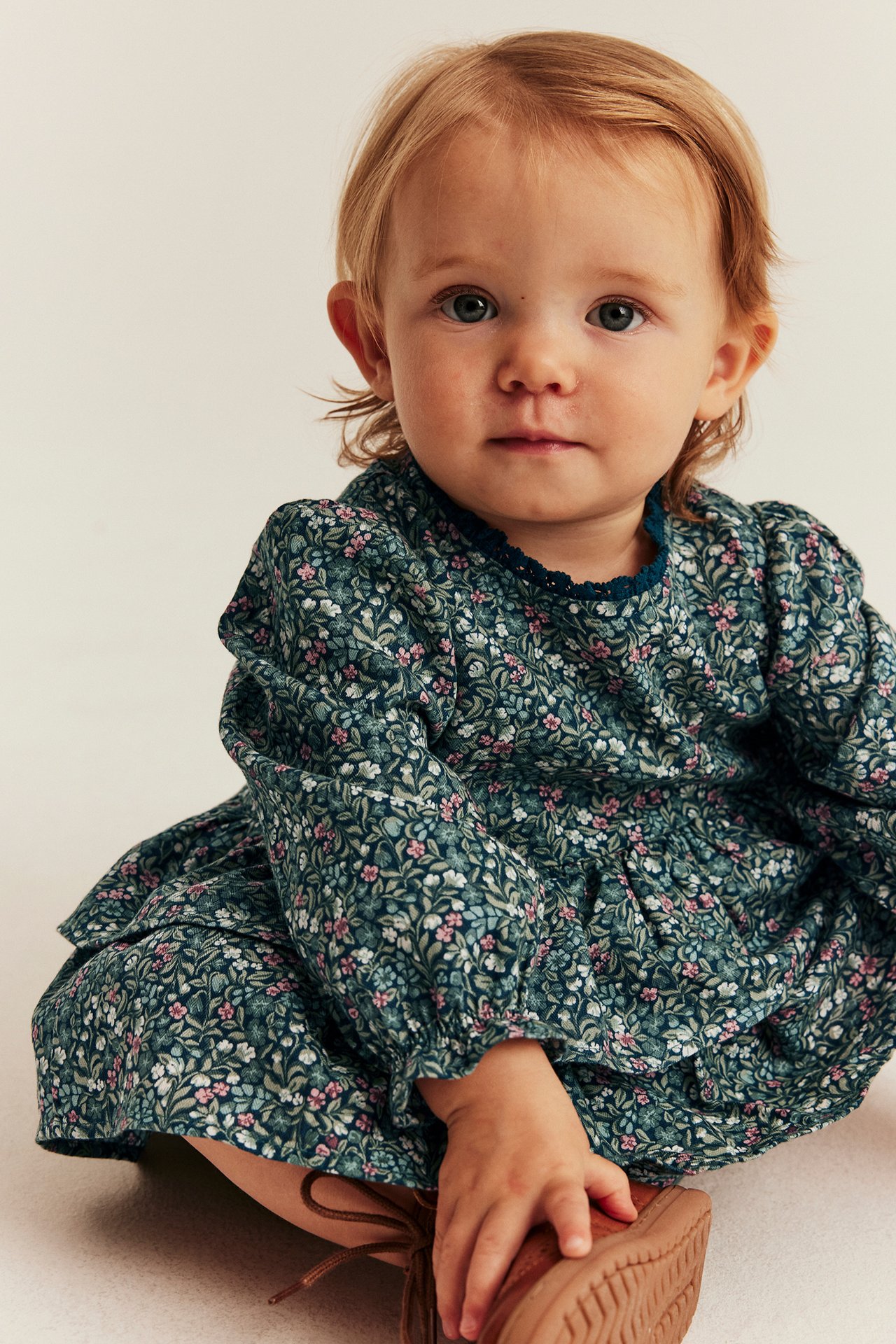 Front view, baby wearing a dark green floral print dress with ruffled collar and brown shoes.