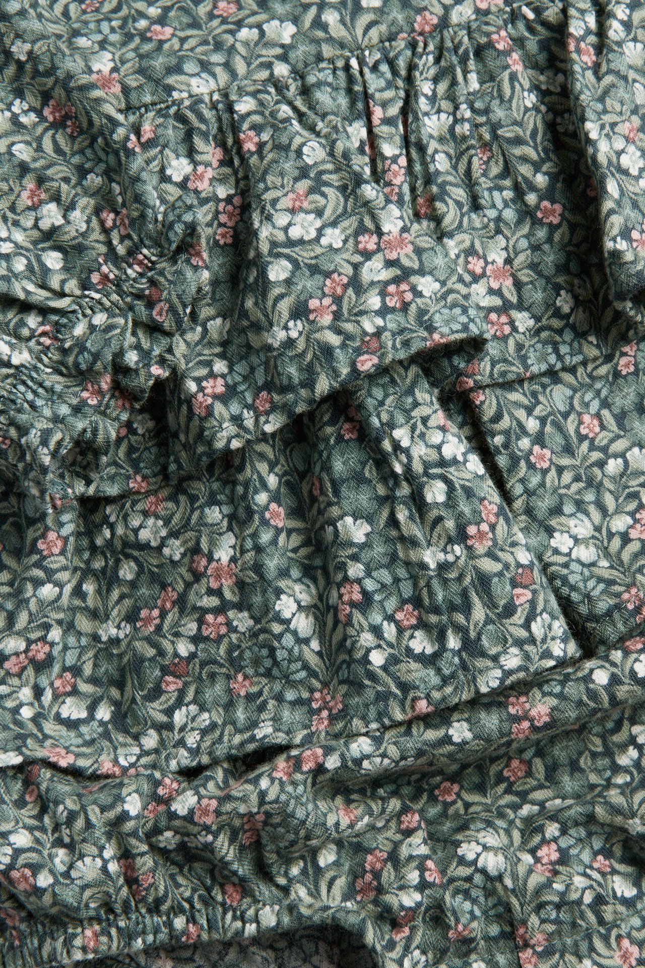 Nahaufnahme eines dunkelgrünen Stoffes mit Blumenmuster, weißen und rosa Blüten, mit Rüschen.