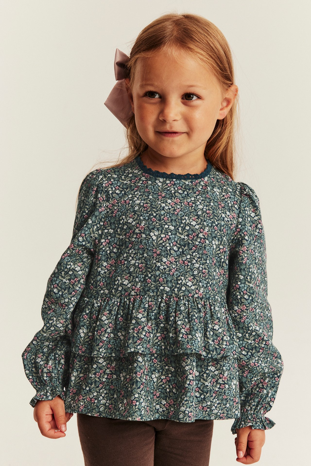 Front view: Kid in dark floral long-sleeved tiered top, brown leggings, and a mauve hair bow.