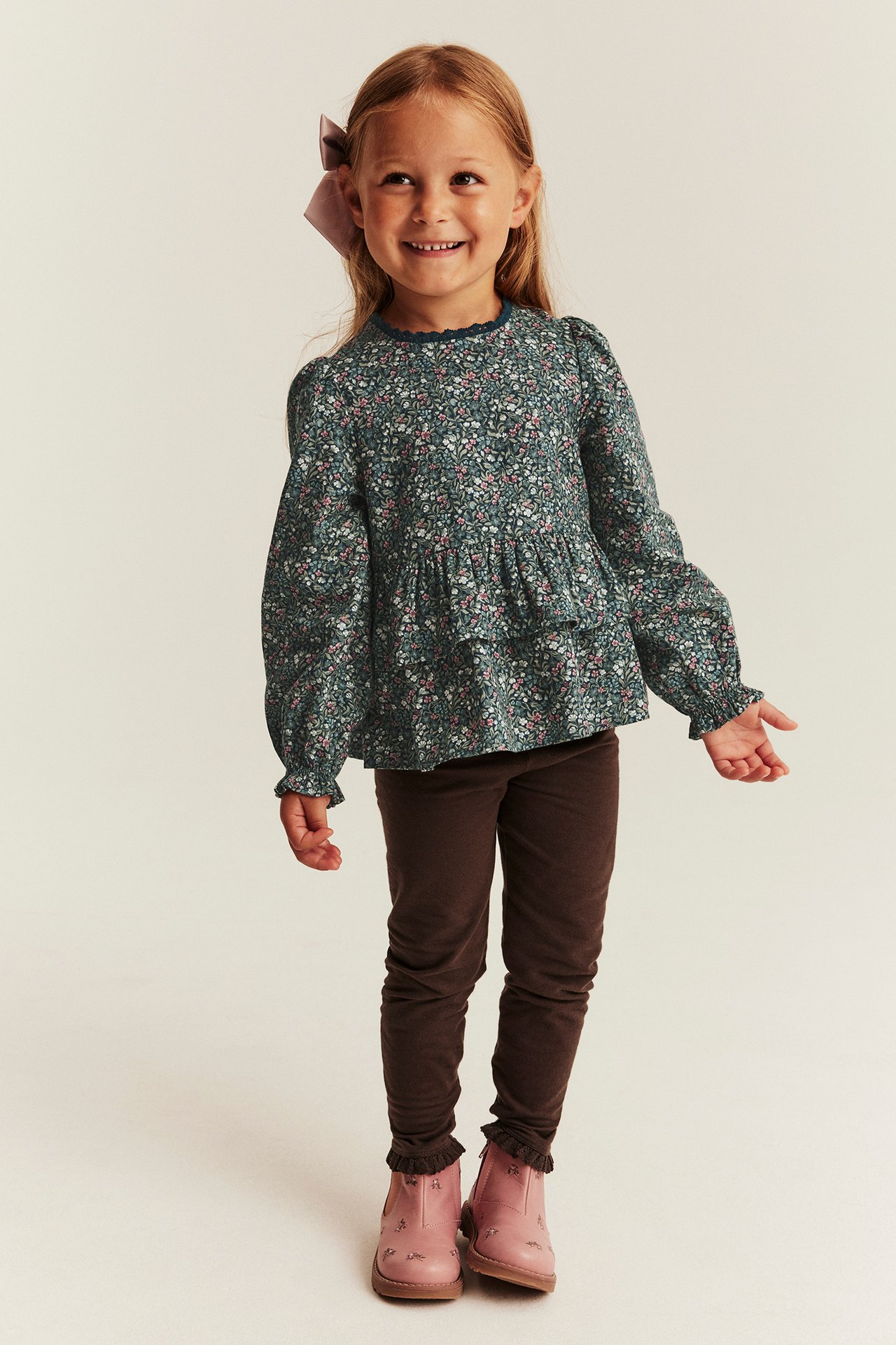 Front view of a girl wearing a green floral long-sleeved peplum top, brown leggings, and pink boots.