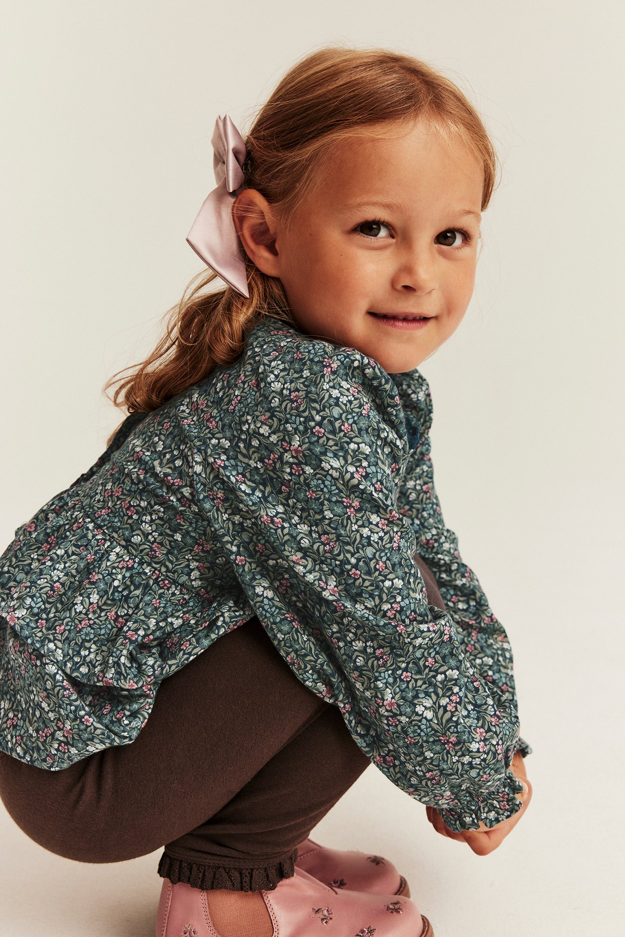 Front view of a kid wearing a dark green floral long-sleeve blouse, brown leggings, and pink shoes.