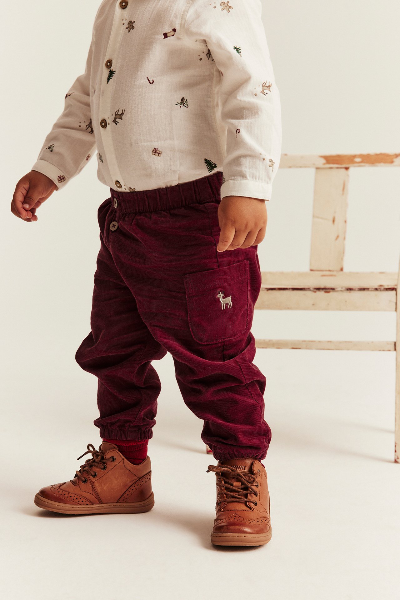 Baby in white Christmas print shirt, burgundy corduroy pants with deer, brown lace-up shoes.
